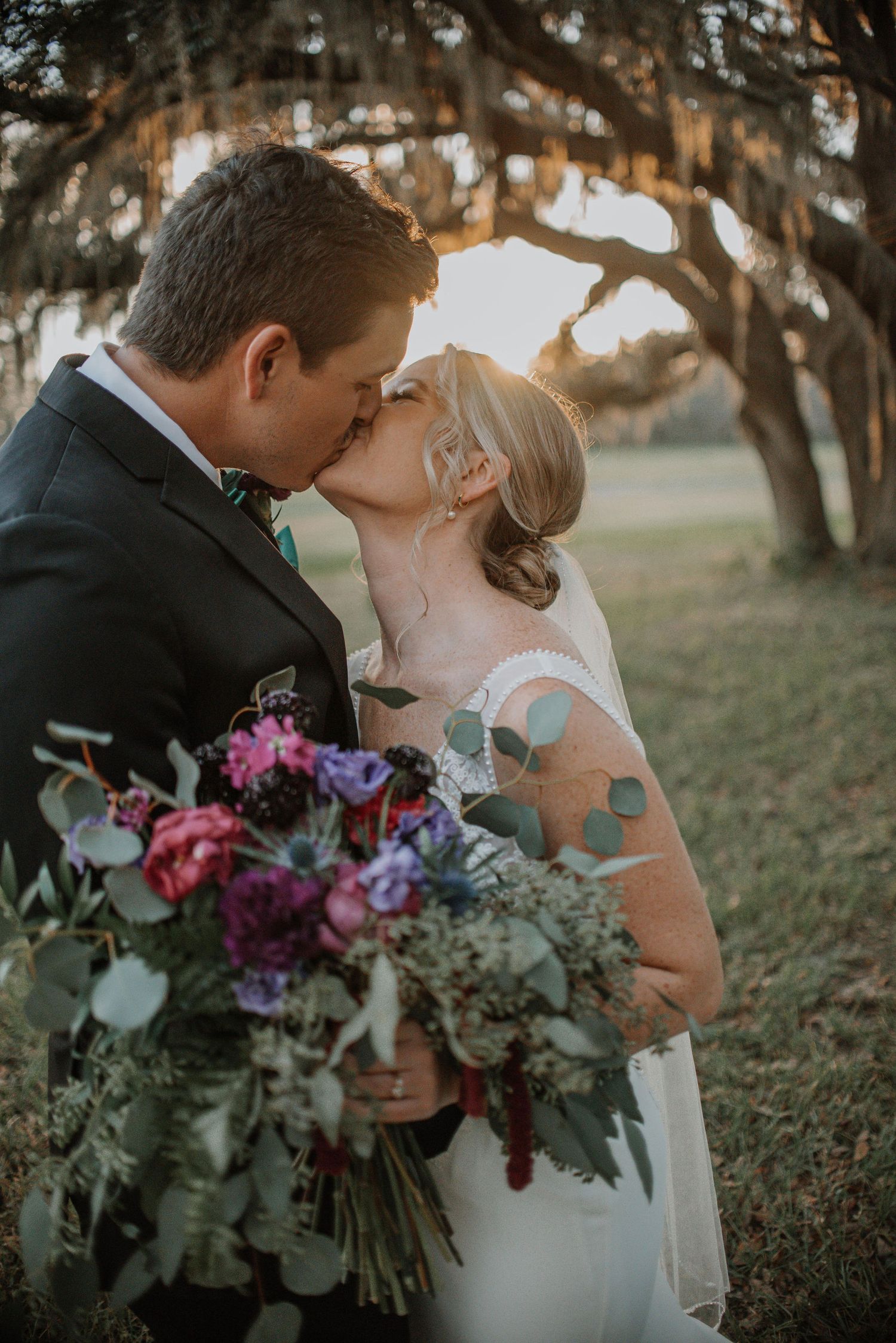 Newlywed couple shares intimate moment with colorful bouquet under trees.