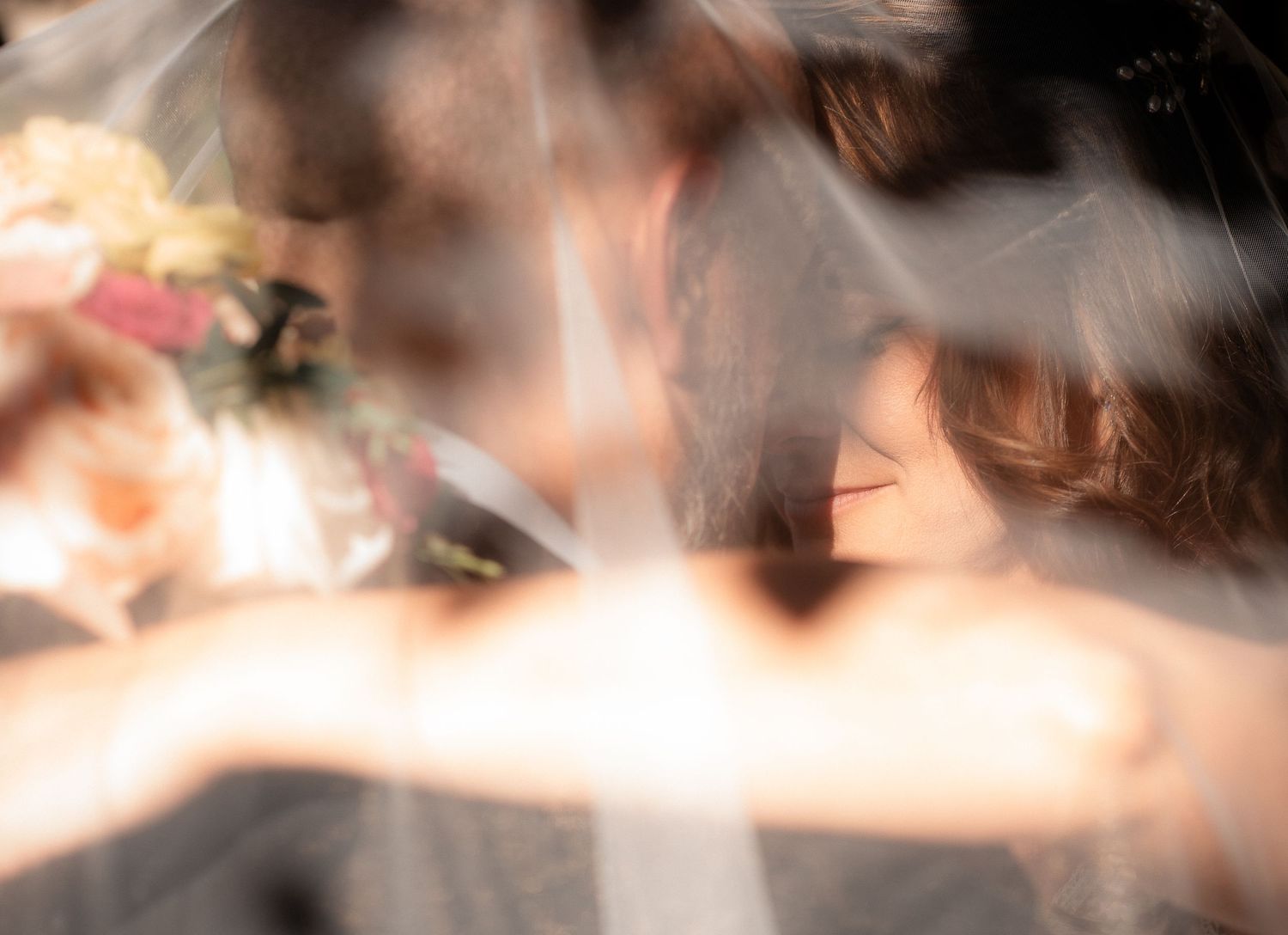 Blurred romantic wedding moment of a couple embracing behind a sheer veil, with a pastel bridal bouquet of pink and peach flowers nearby.