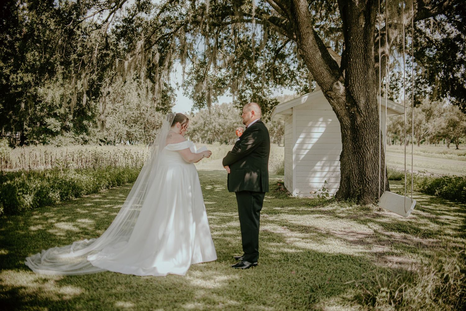 Bride in a long white gown and veil faces the groom in a dark suit during an outdoor wedding ceremony beneath a large moss-draped oak tree.