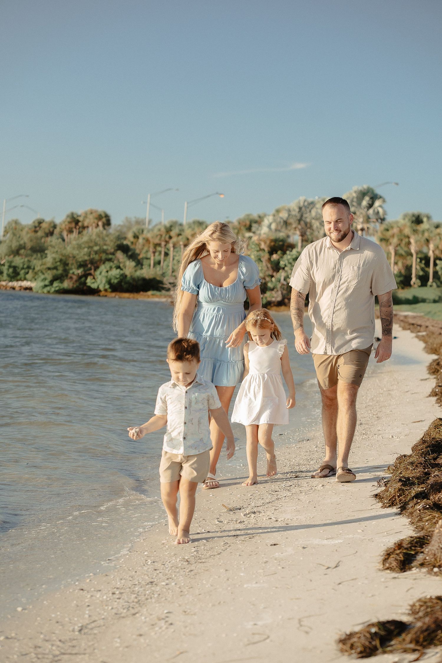 Family walking together along sunny beach shoreline in light summer clothing.