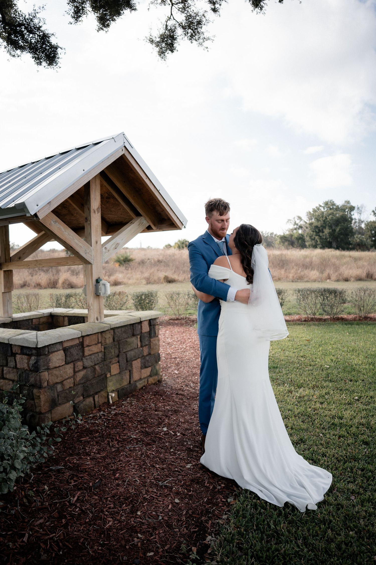 Bride and groom share a romantic embrace near a rustic stone well in an outdoor countryside wedding setting.