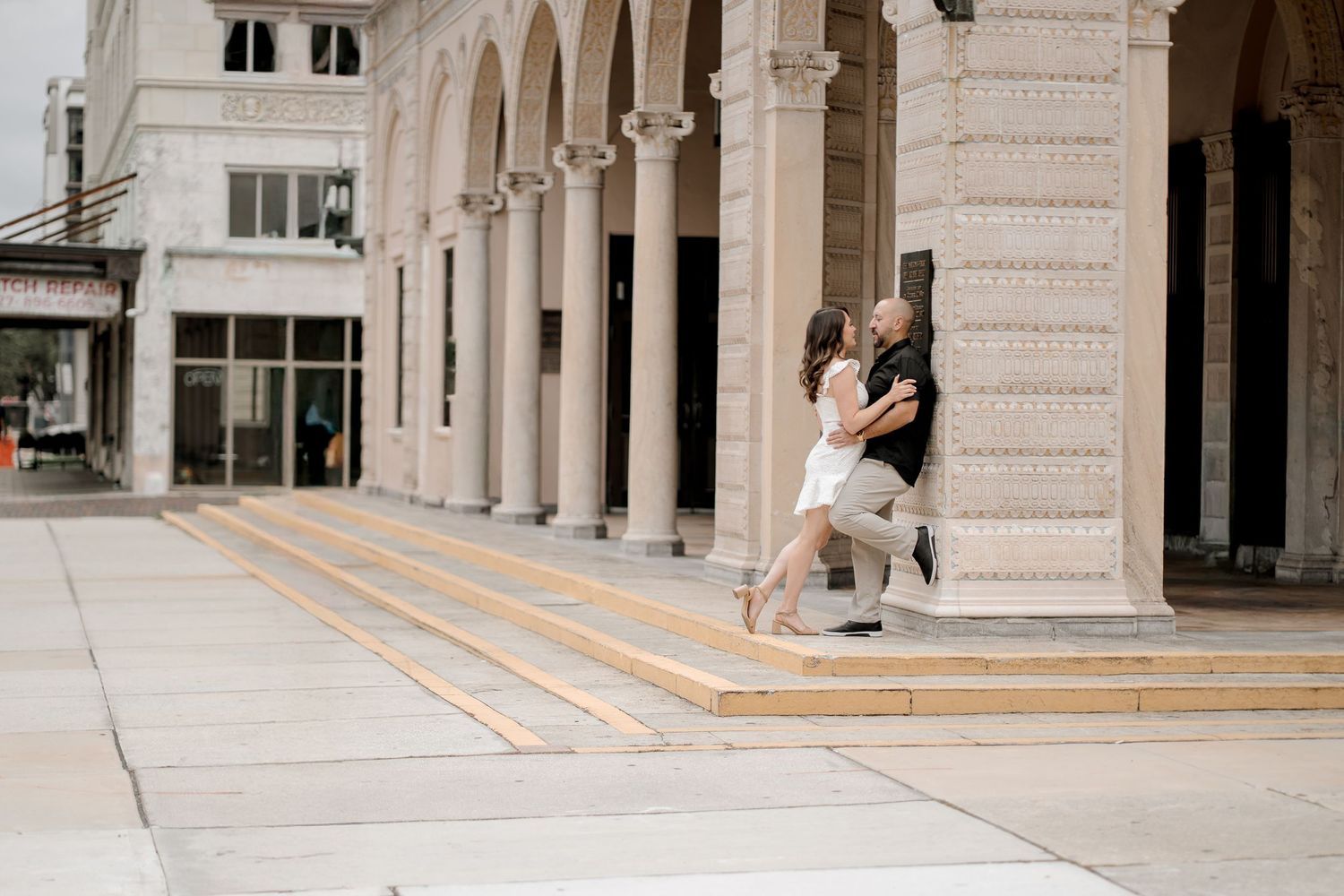 A couple shares a romantic moment under classical stone archways of an elegant building.
