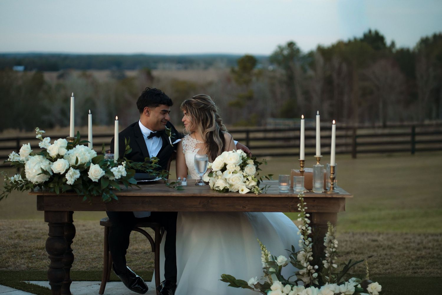 Bride and groom share romantic moment at floral sweetheart table with candles at outdoor wedding reception.