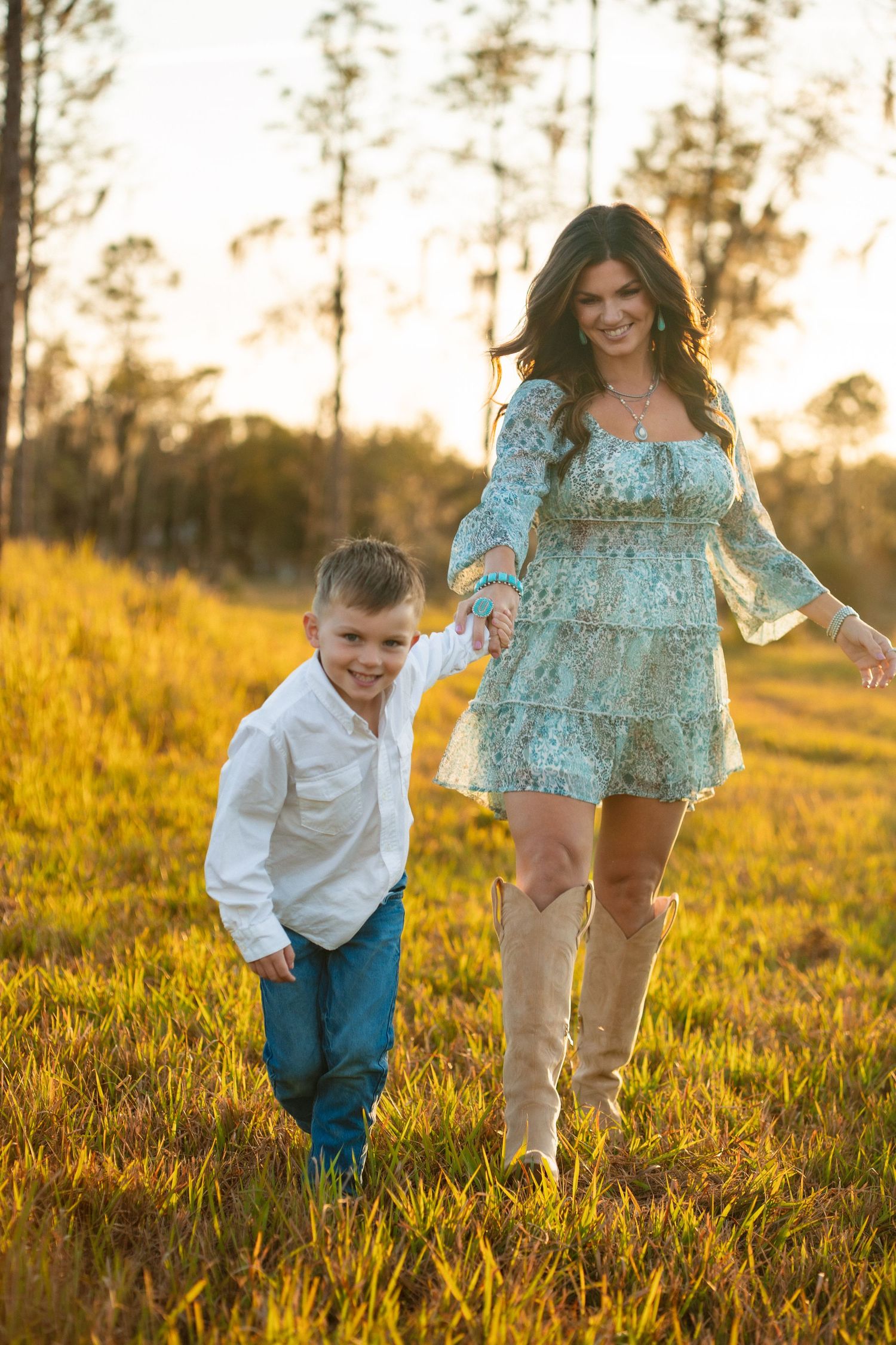 A child and woman run playfully through a golden field at sunset while wearing blue and white outfits.