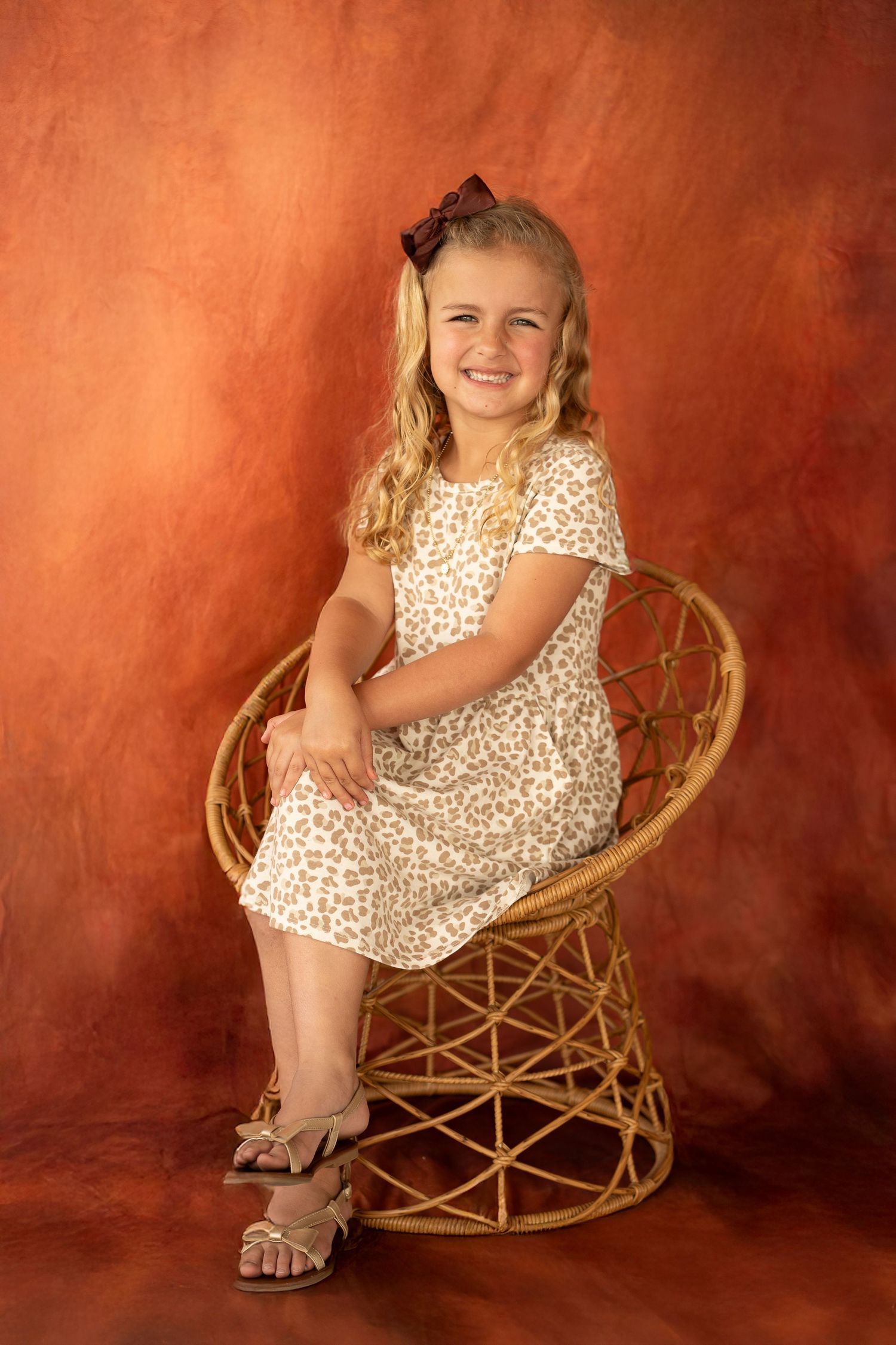 A child sits in a woven peacock chair wearing a polka dot dress against a warm orange-toned backdrop.