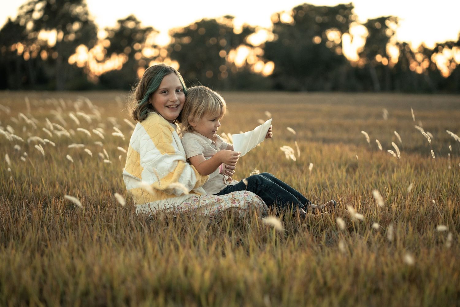 Two people sit together in a golden field of tall grass at sunset reading a book together.