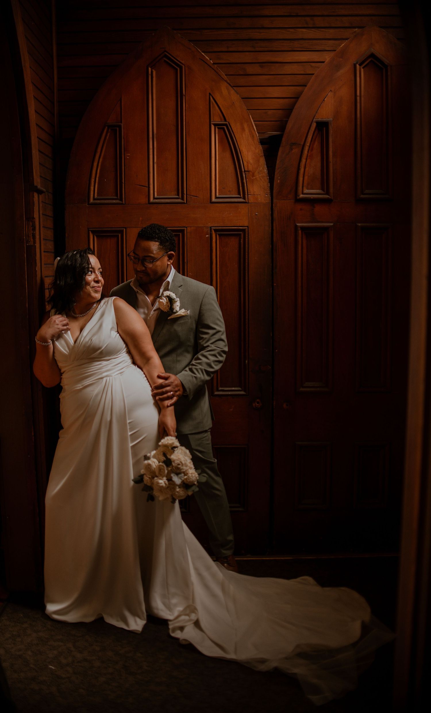 A couple poses elegantly against wooden church doors in dramatic lighting.