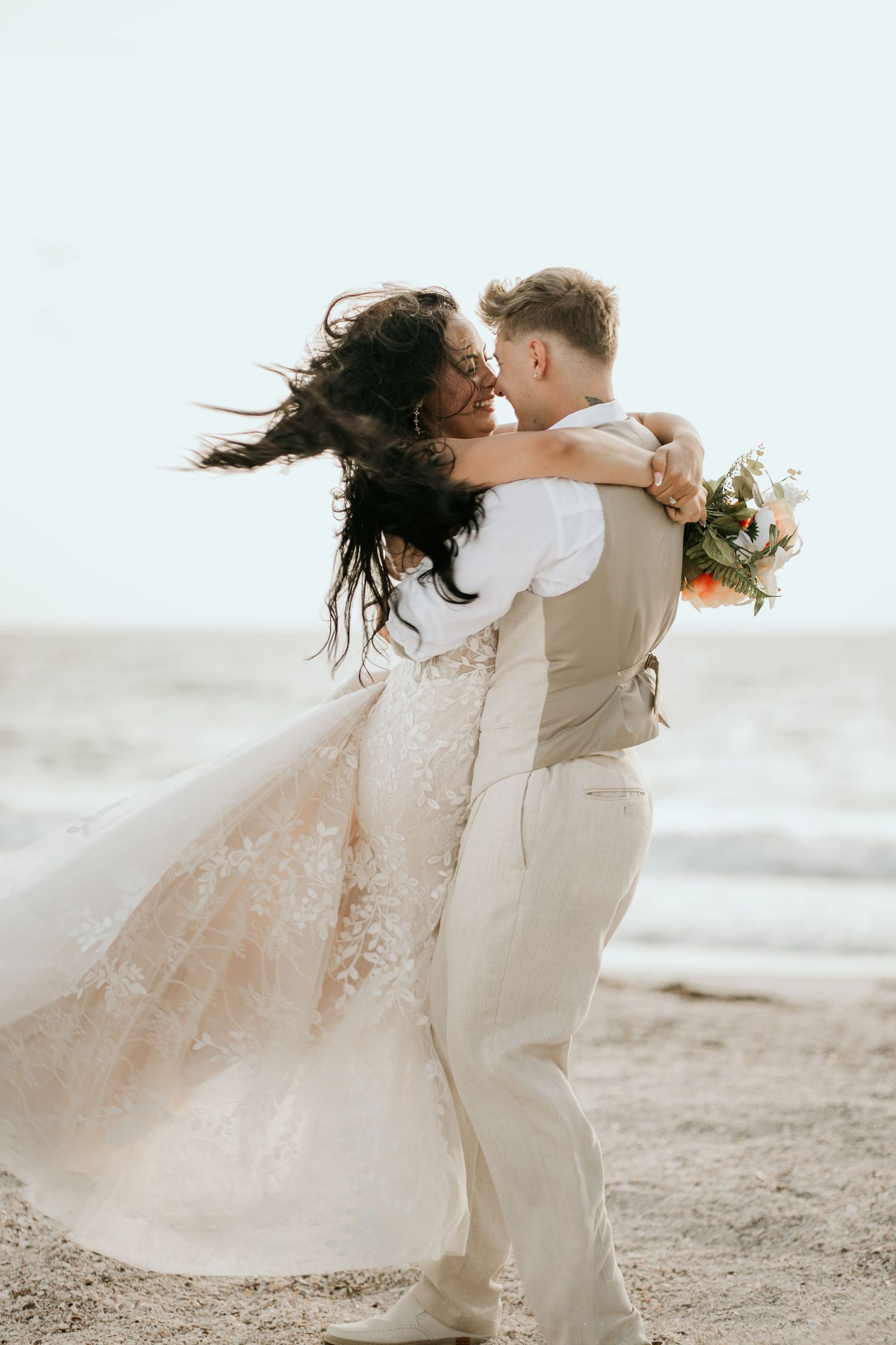 A romantic beach wedding photo of a couple embracing with flowing dress and beige suit against an ocean backdrop.