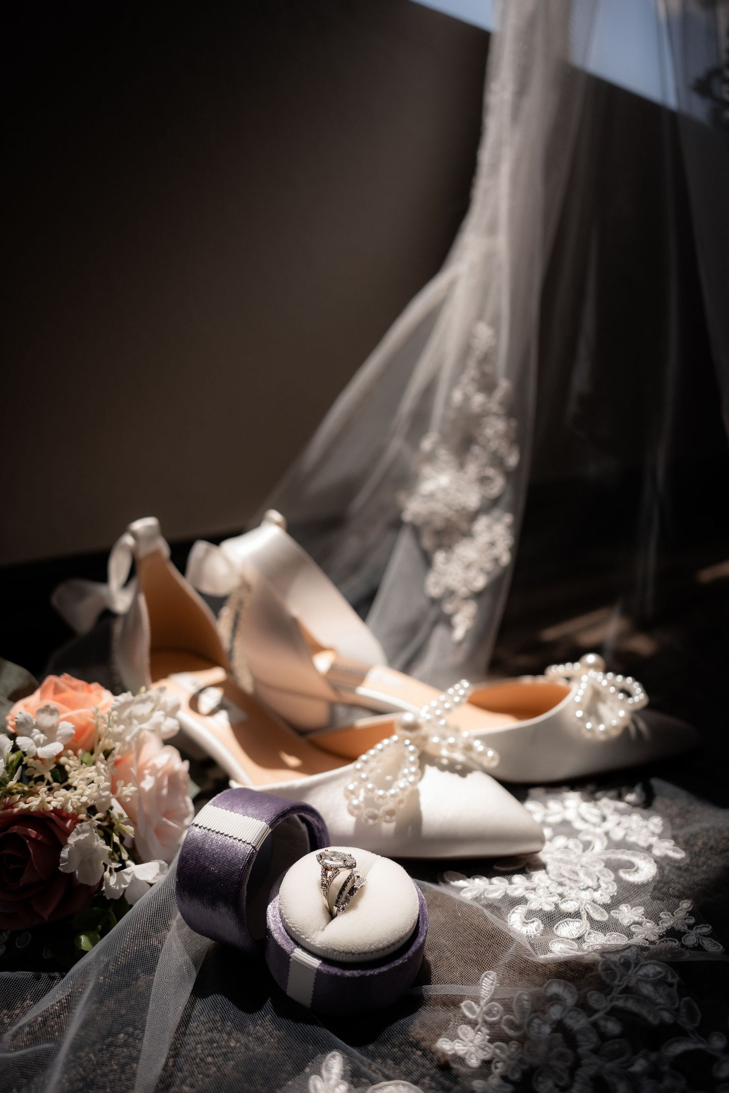 White bridal heels with pearl details, diamond engagement ring in purple box, and floral bouquet on lace veil.