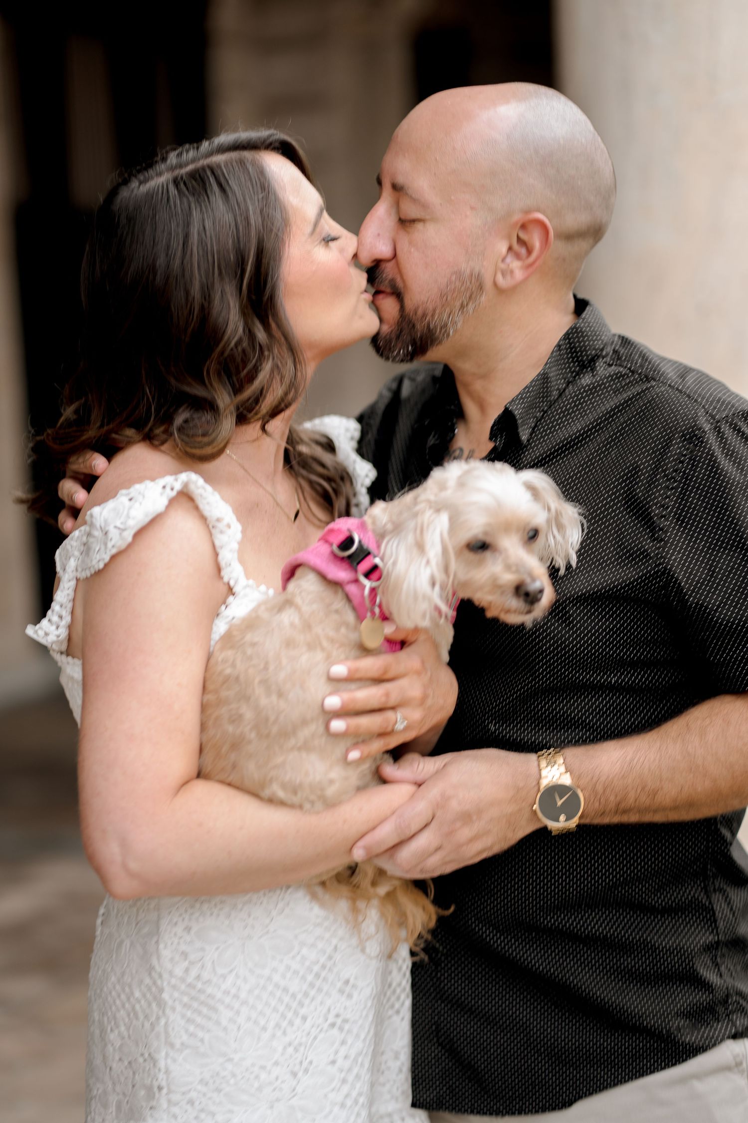 Couple sharing romantic moment while holding small white dog in formal attire.