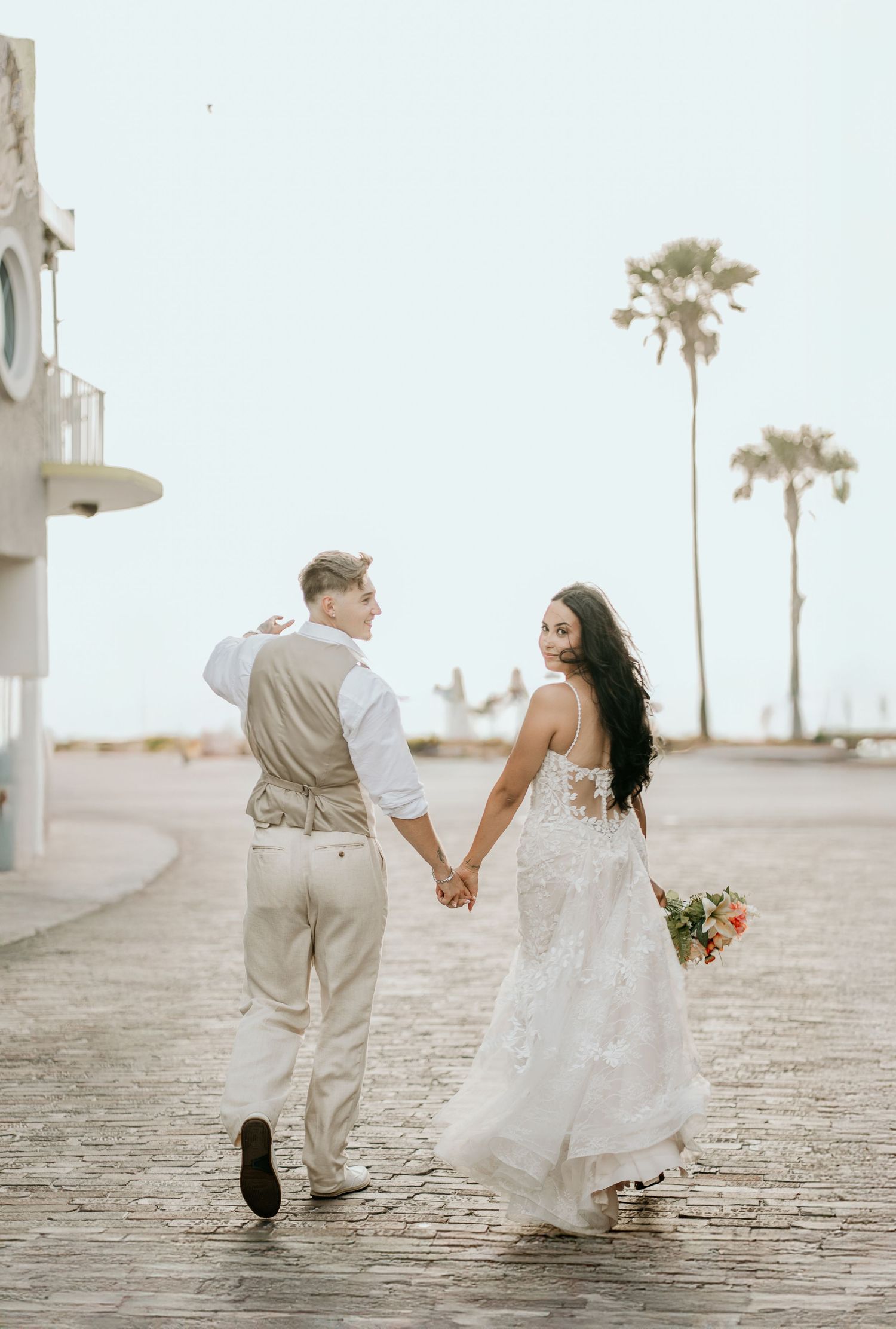 A couple walks hand in hand along a beachfront path with palm trees in the background.