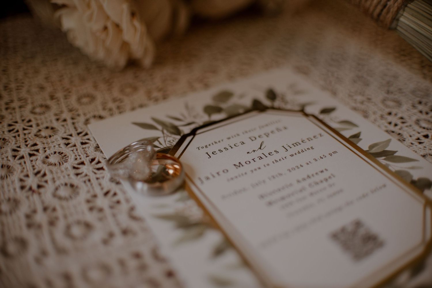Elegant wedding invitation with gold-trimmed botanical border and QR code resting on lace fabric beside a sparkling diamond engagement ring.