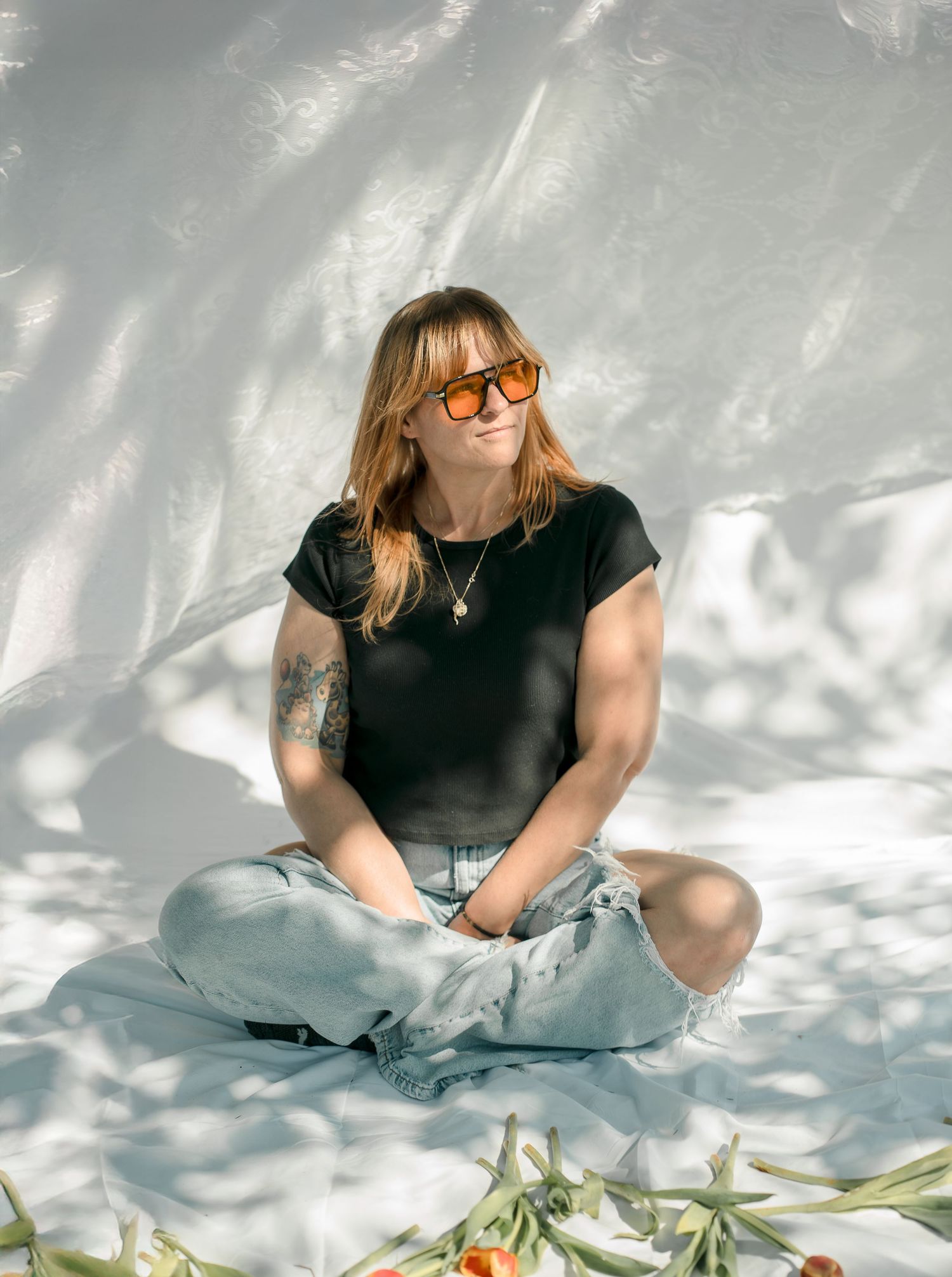 Portrait of woman in black shirt and jeans sitting cross-legged with orange flowers.