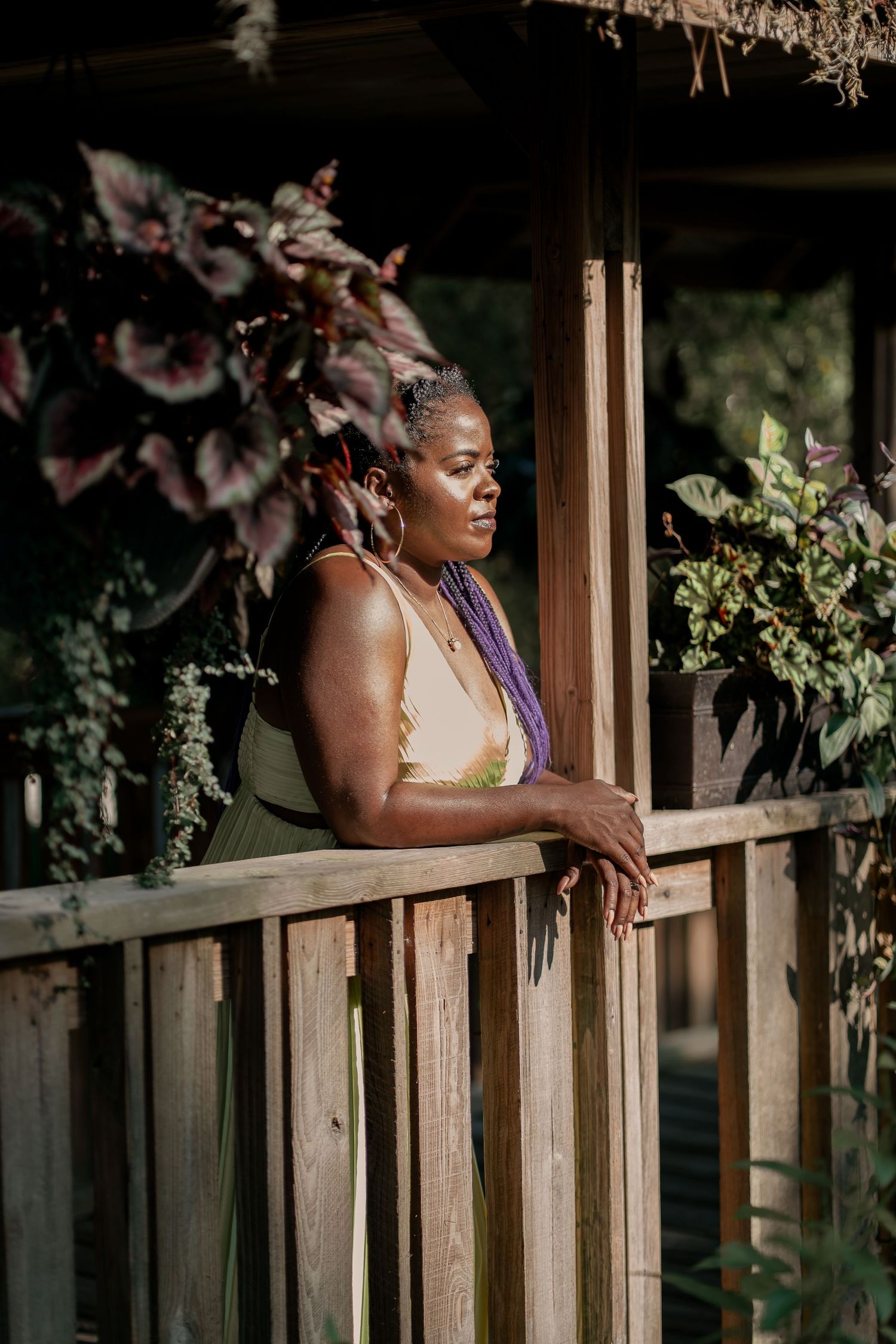 Woman in yellow dress leans on wooden railing surrounded by lush garden foliage.