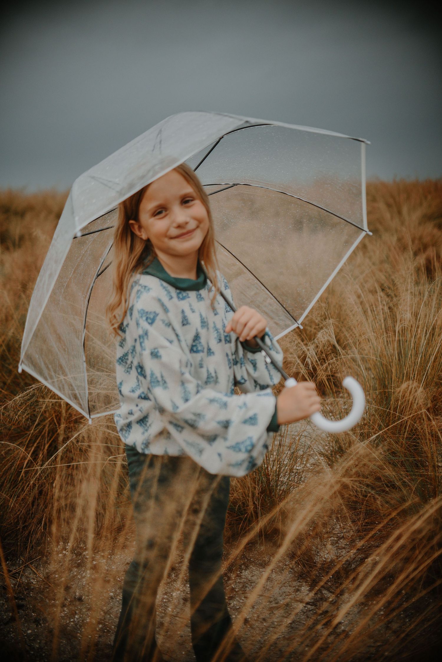 Child holding clear umbrella poses playfully in tall grass field at sunset.