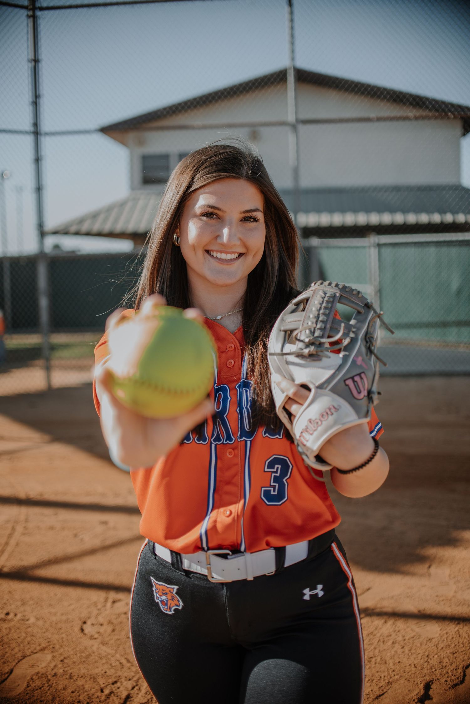 Softball player in orange jersey holding glove and ball on dirt field.