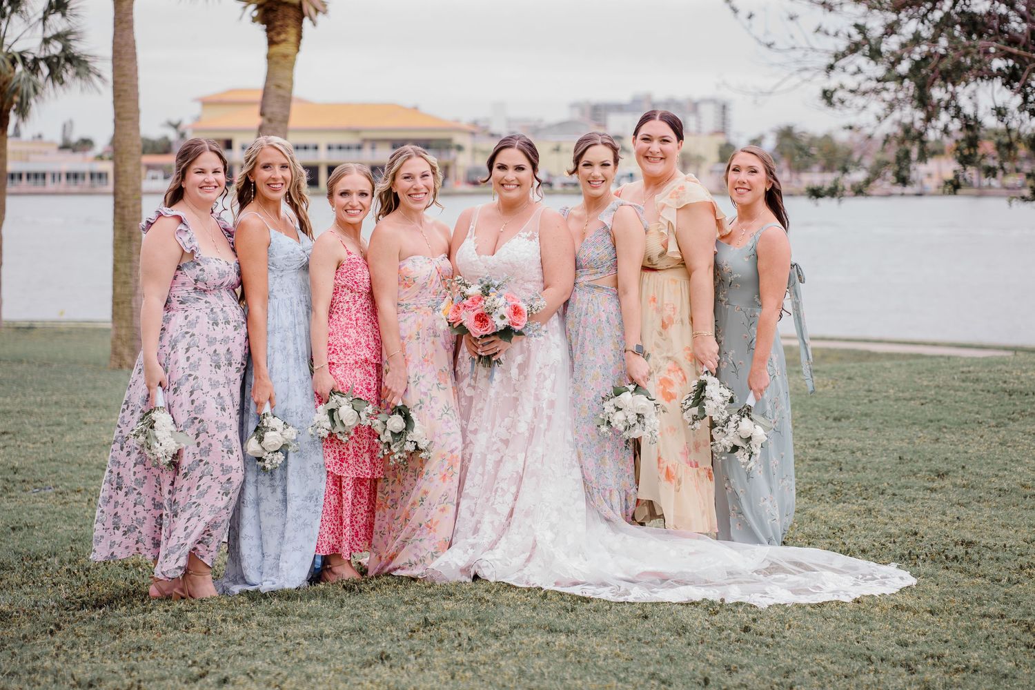 A bridal party in pastel floor-length dresses poses together on a grassy lawn near palm trees.