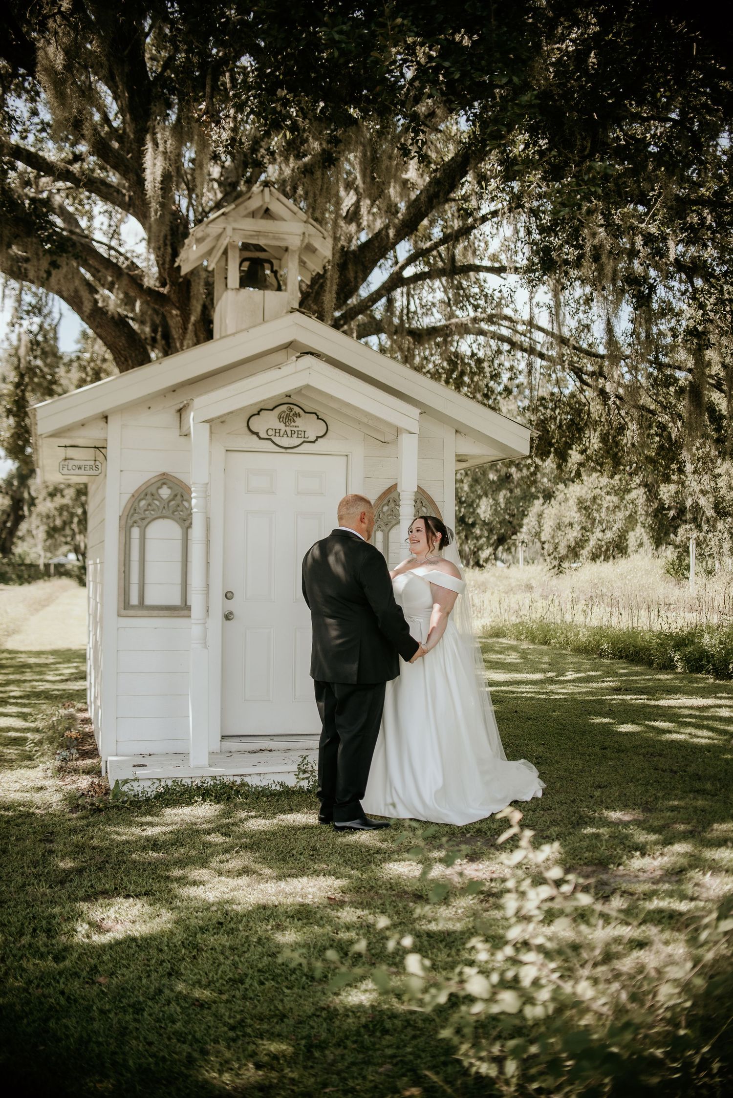 Wedding couple shares tender moments near white chapel under Spanish moss trees.