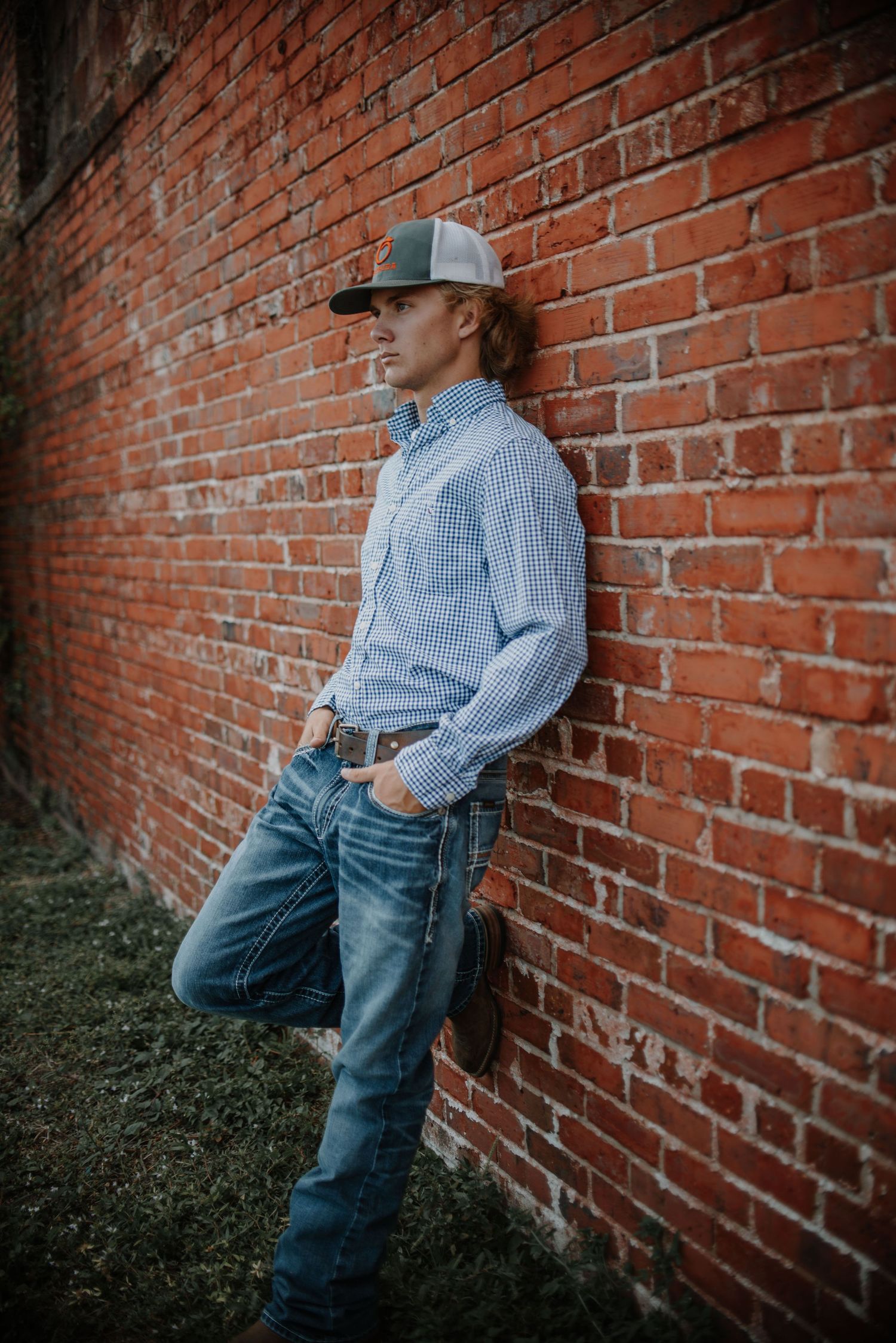 Artistic portrait against rustic brick wall wearing baseball cap and casual attire.