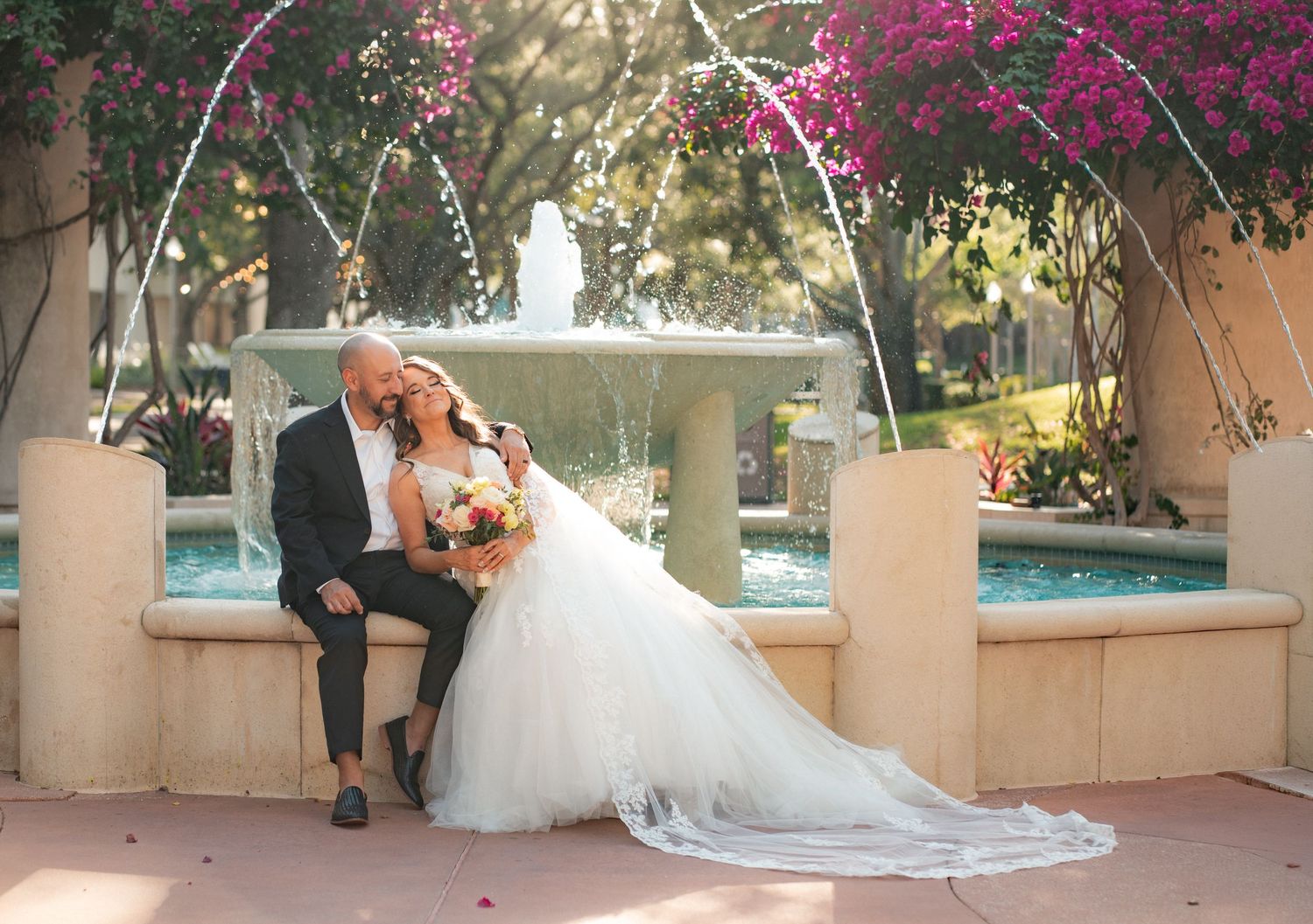 Bride in a white gown with a long train and groom in a dark suit sit close on a stone fountain ledge beside blooming pink bougainvillea.