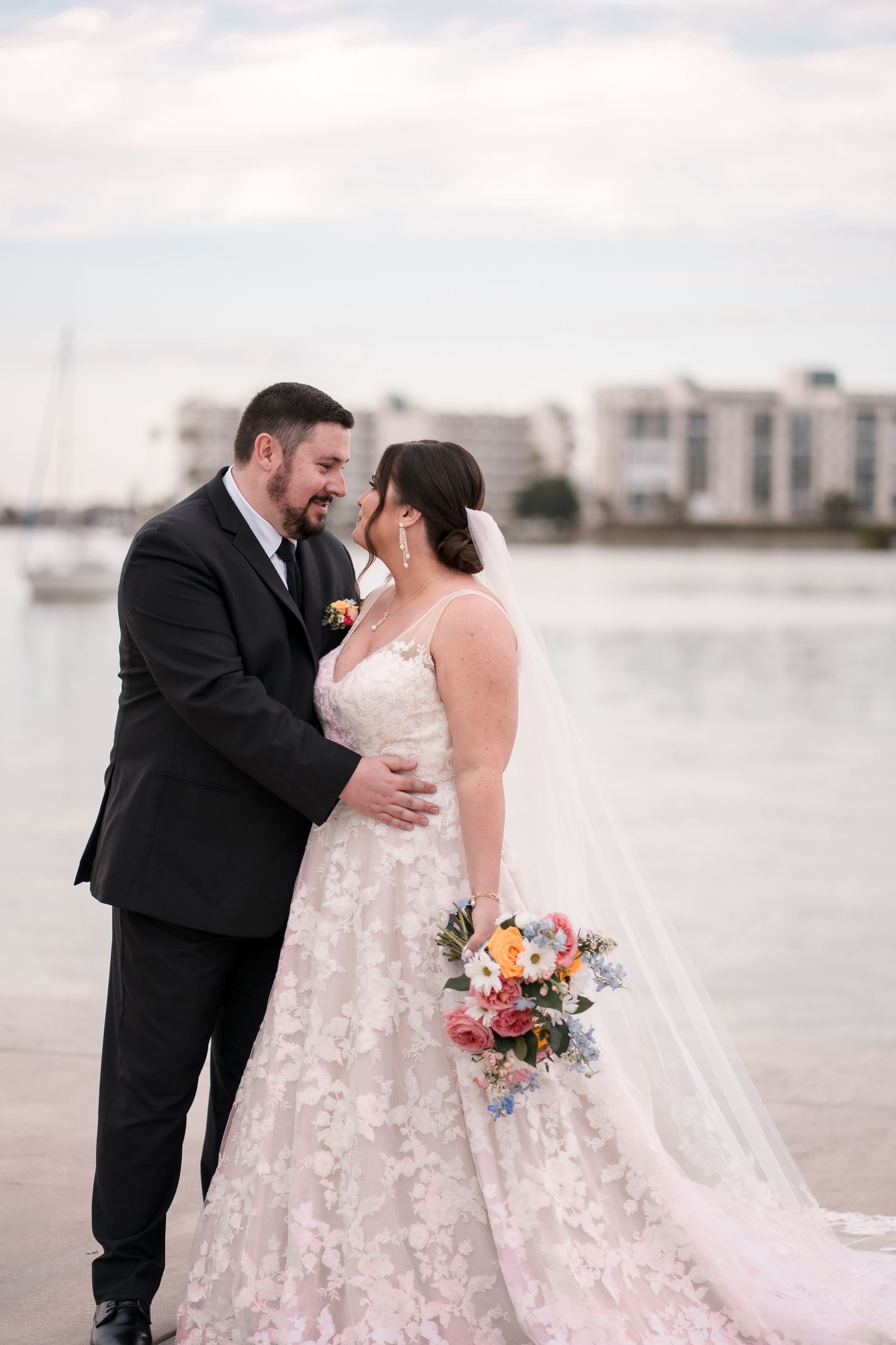 Bride in a lace gown with a long veil holding a colorful bouquet embraces her groom in a black suit beside a waterfront at dusk.