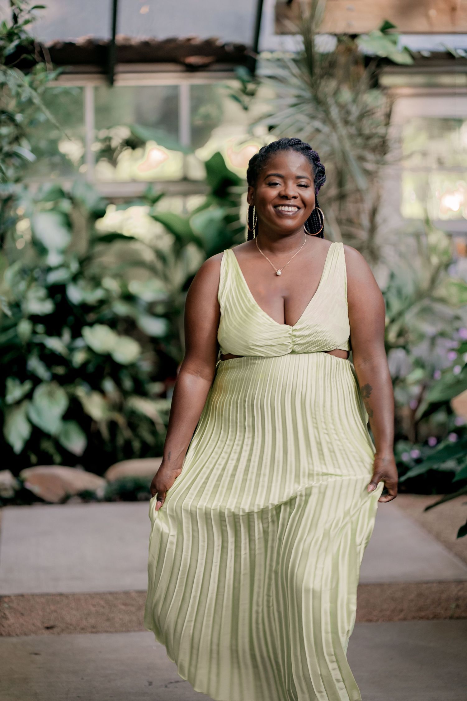 Woman in flowing pale yellow pleated dress poses in a greenhouse setting.