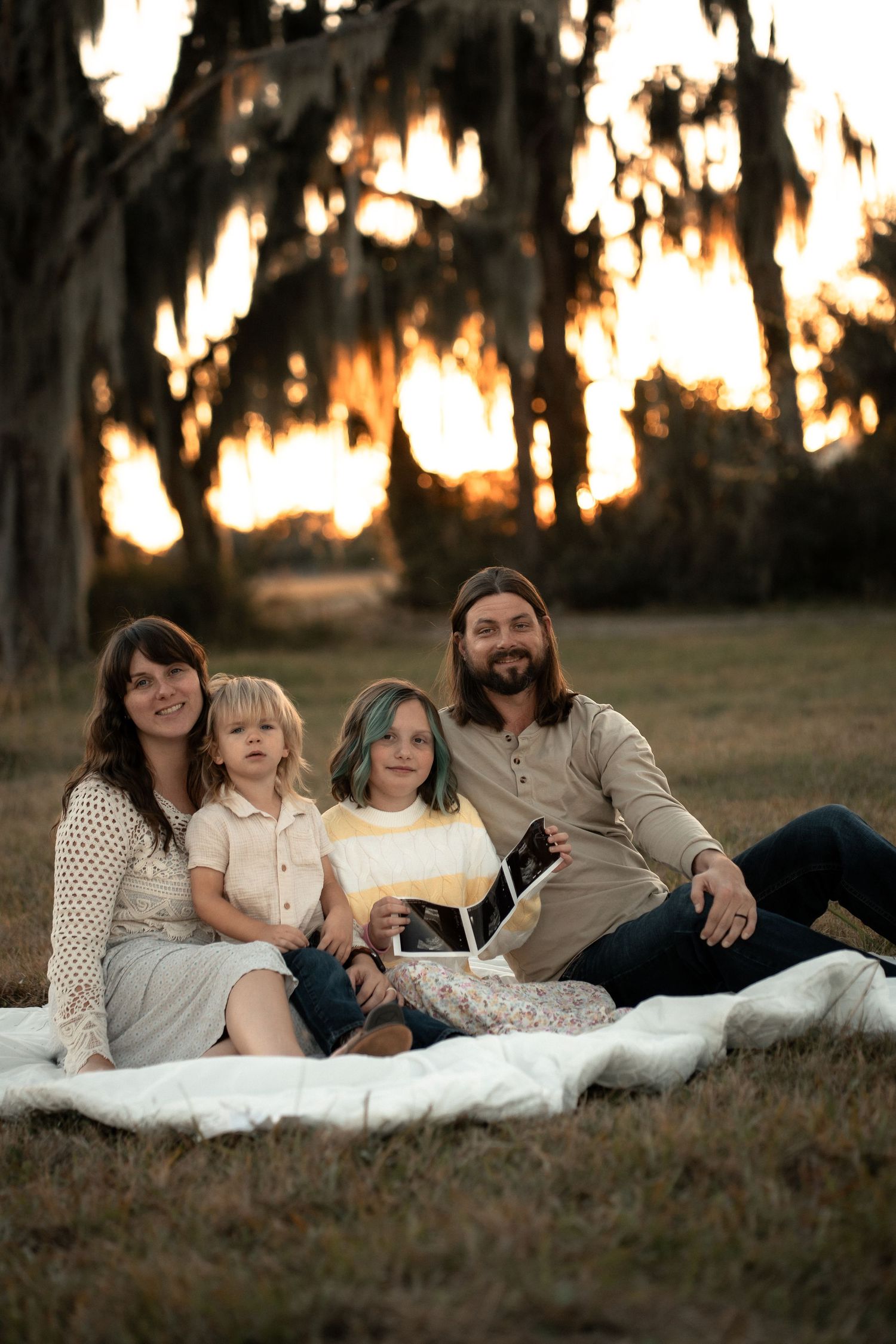 A group relaxes together on a white blanket in a grassy field at sunset with Spanish moss trees in the background.