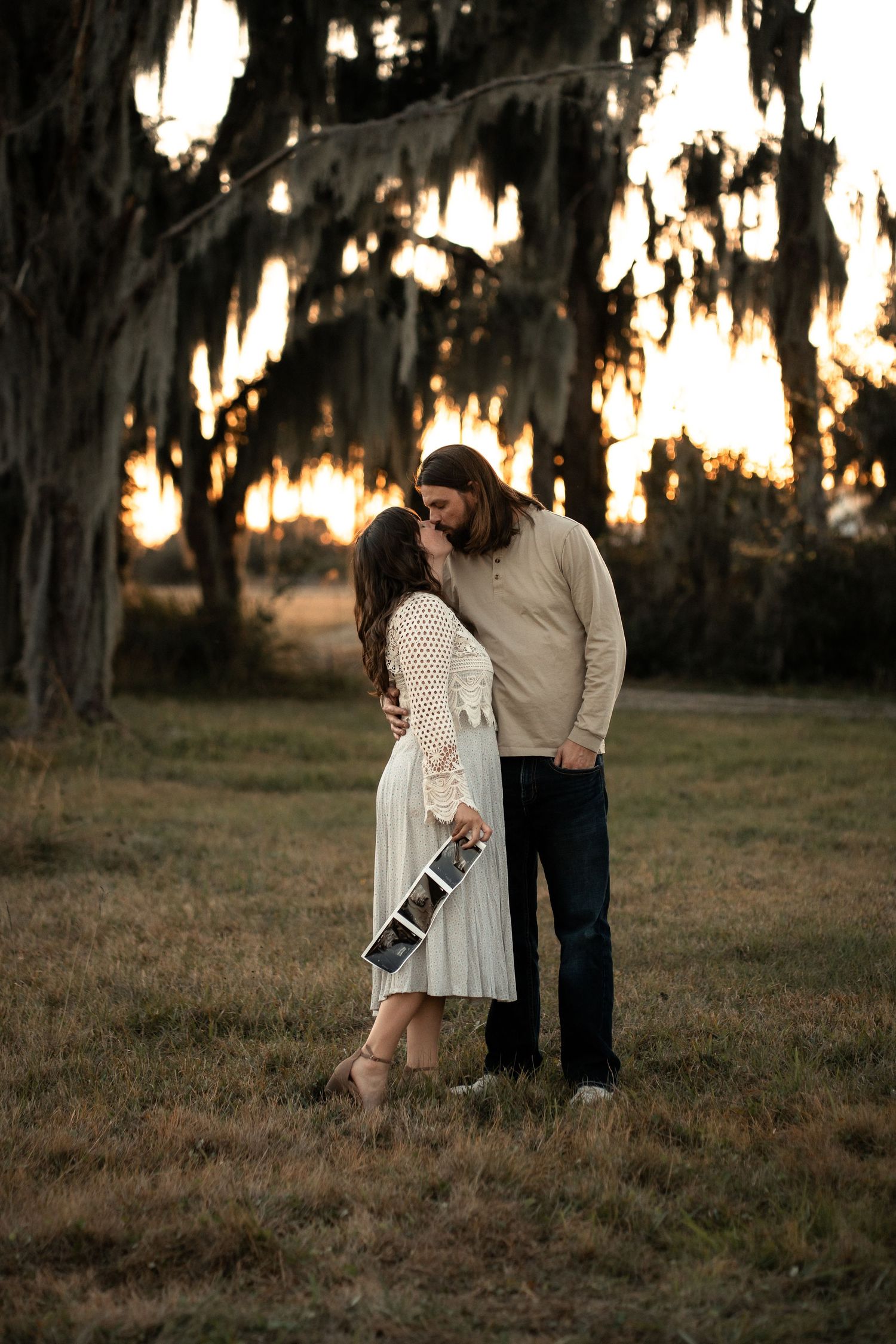 A romantic couple embraces under Spanish moss at sunset in a dreamy outdoor setting while holding a camera.