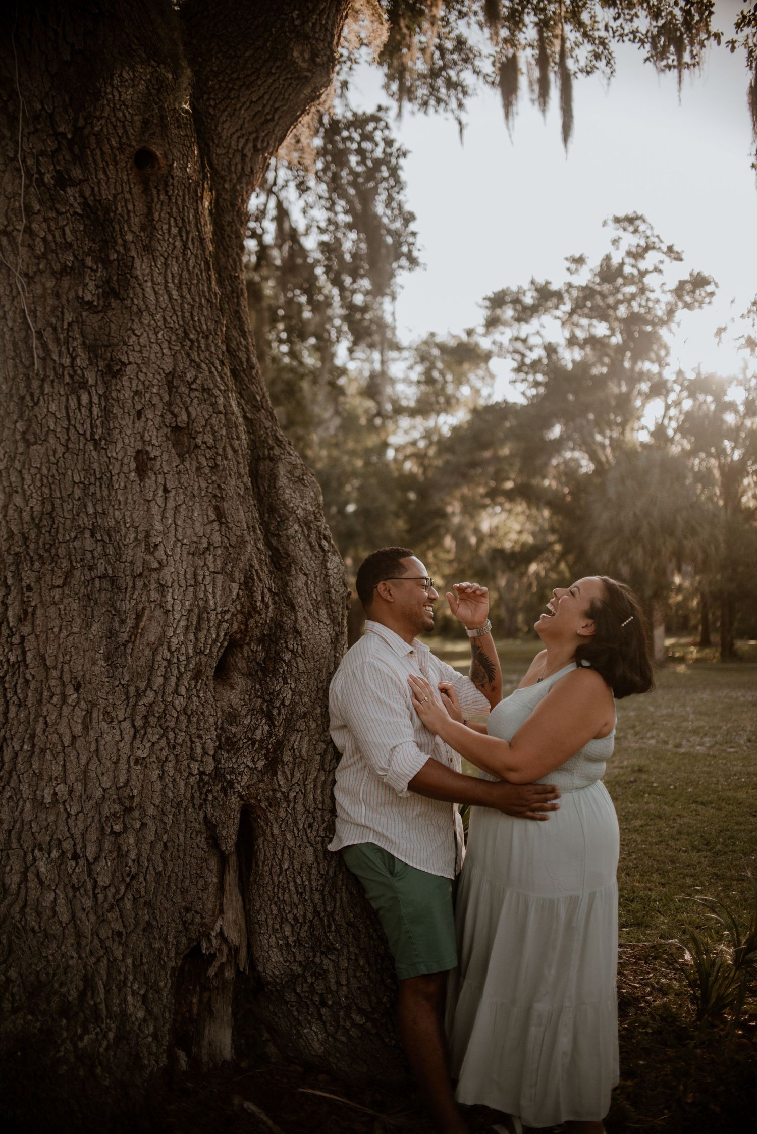 A couple shares joyful laughter beneath large oak trees at sunset.