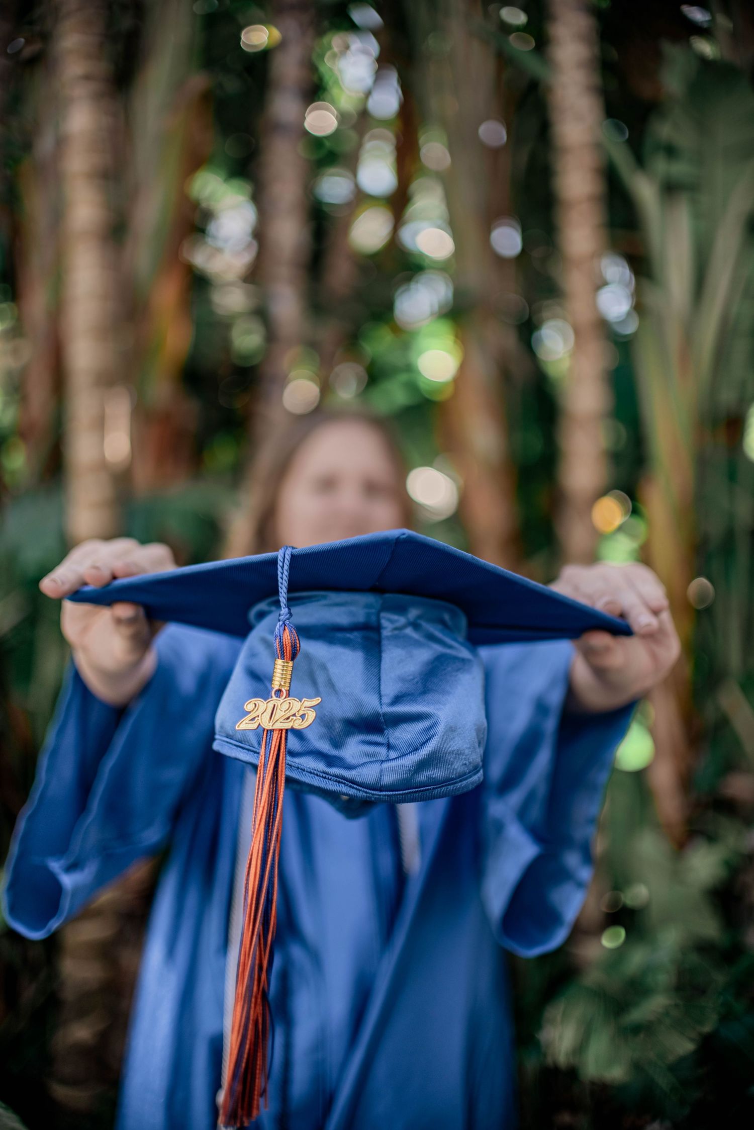 A graduate holds up a blue 2025 graduation cap with tassel outdoors among lush green trees.
