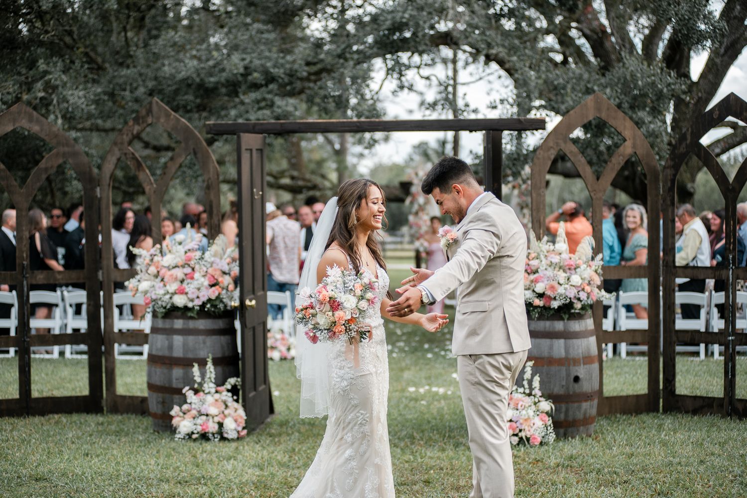 Outdoor wedding celebration with guests in formal attire under large oak trees.