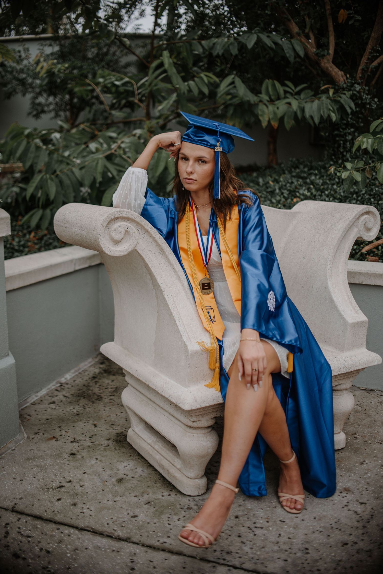 Young woman in blue and gold graduation cap and gown seated on ornate stone bench outdoors.