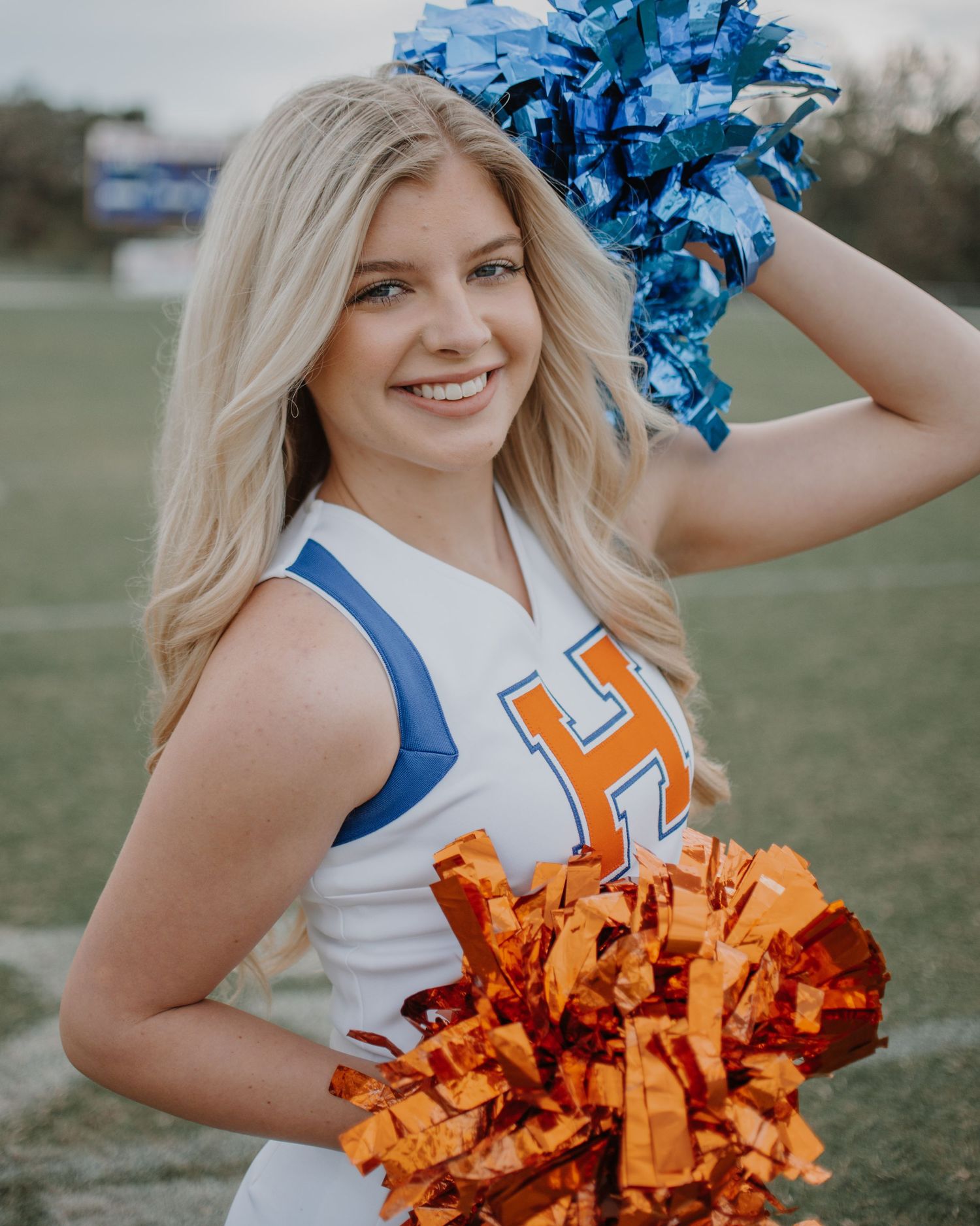 Cheerleader in blue and white uniform waving orange pom poms during performance.
