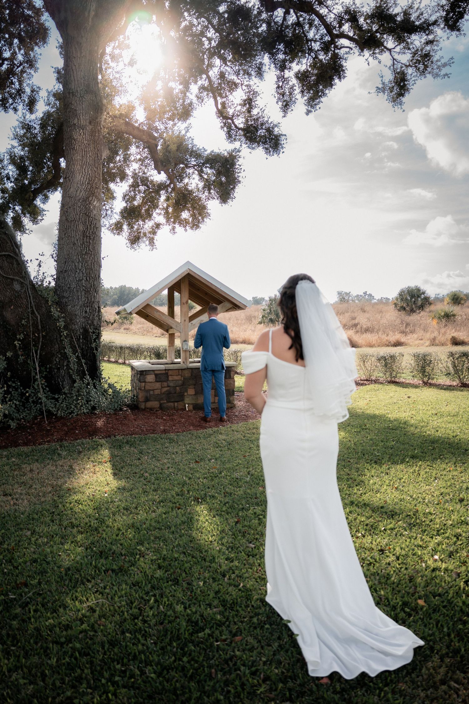 Bride in white gown and veil approaching groom at outdoor wooden gazebo during golden hour wedding ceremony.