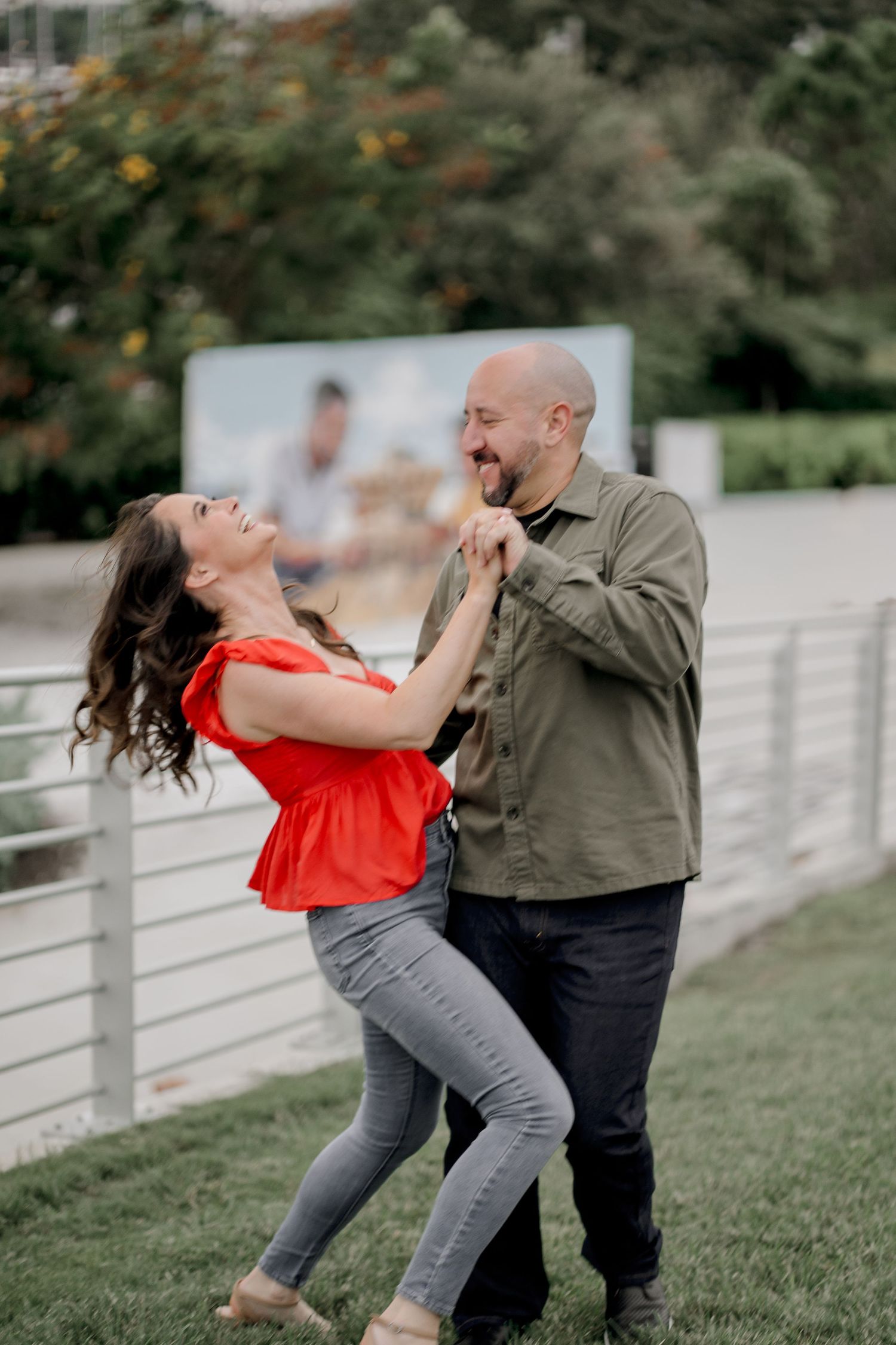 A couple dances and laughs together on grass near a white fence.