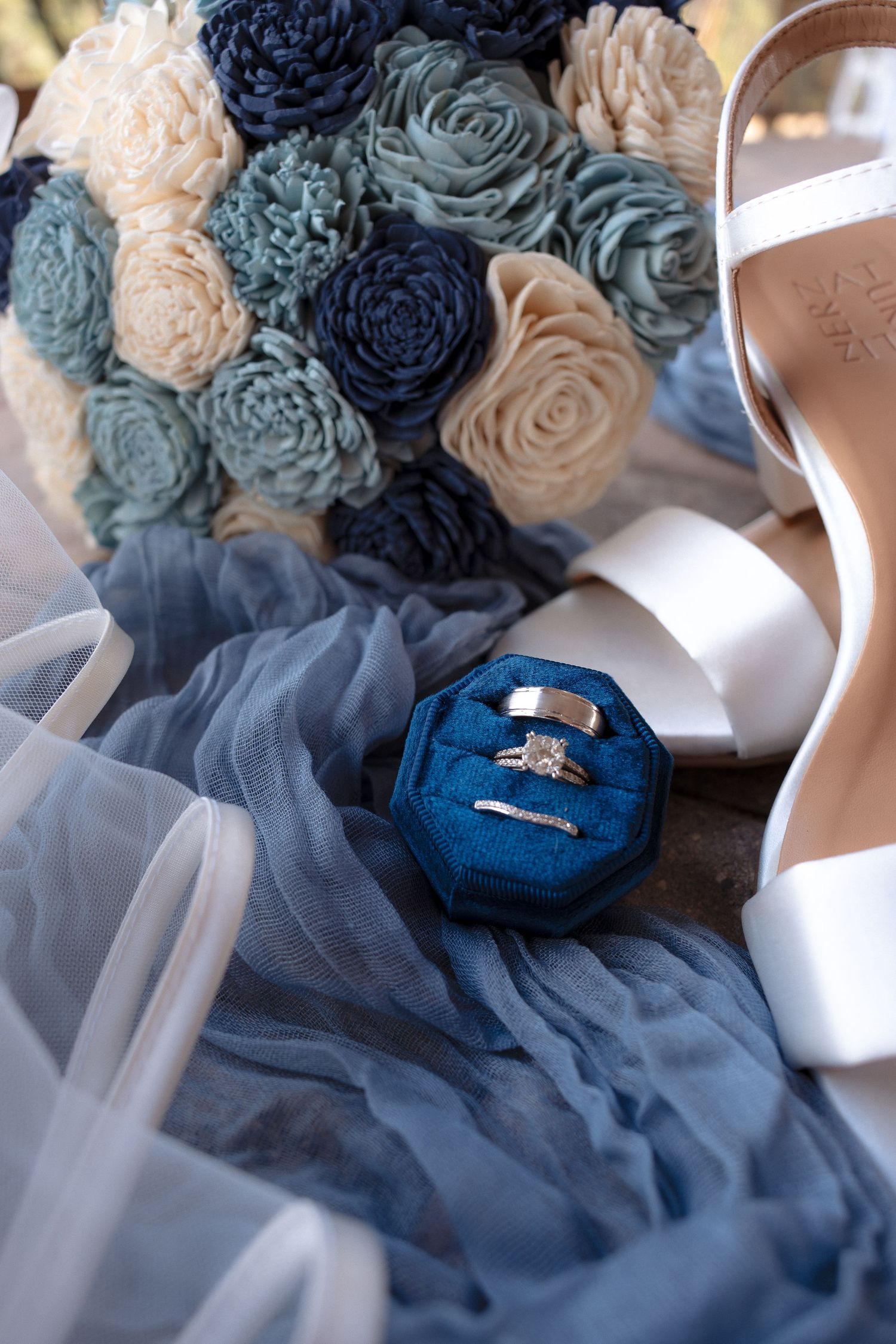 Wedding rings in a blue velvet box beside a dusty blue and cream wooden flower bouquet, white bridal heels, and a flowing veil.