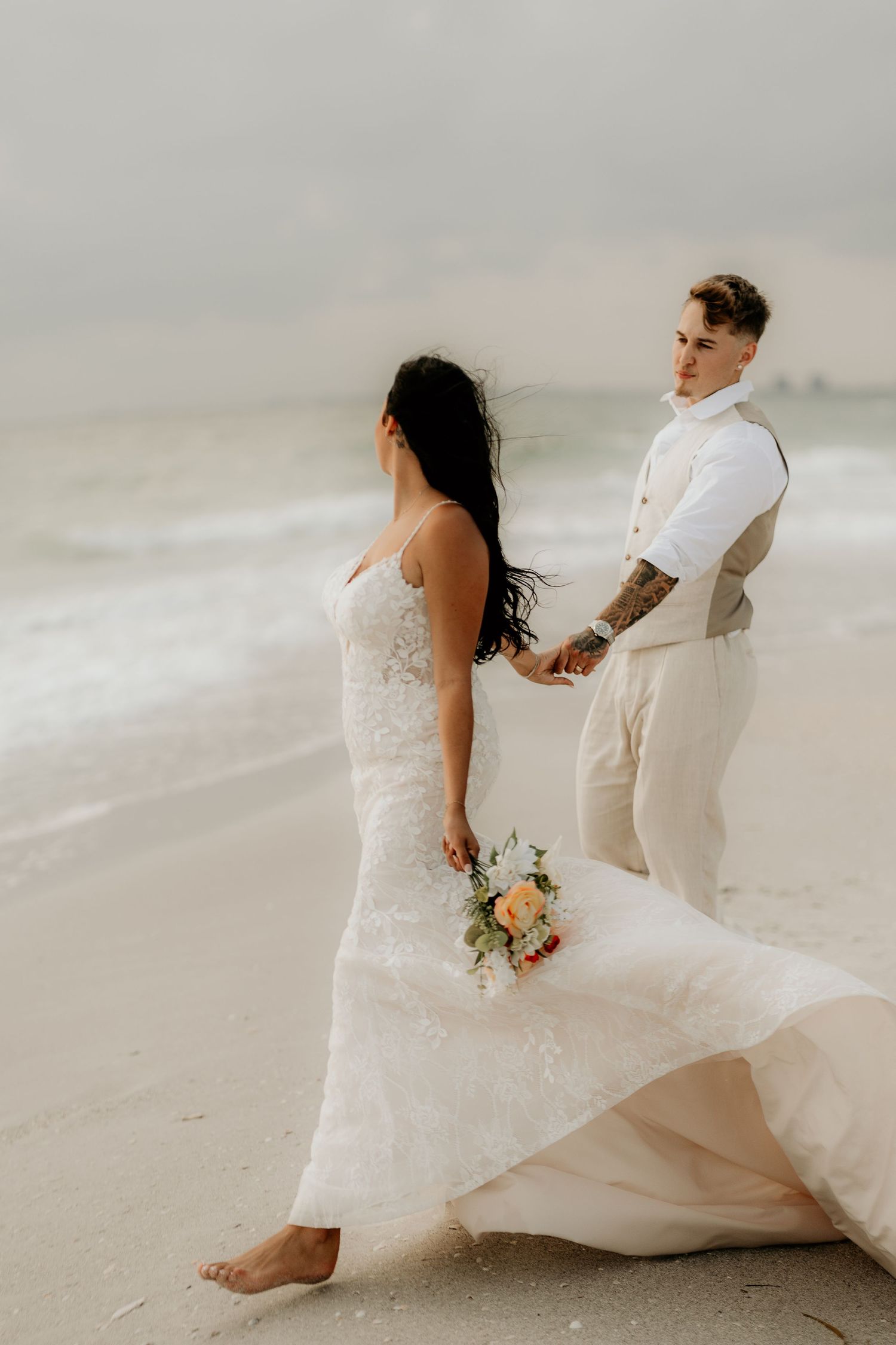A beach wedding couple walking hand in hand along the shoreline in elegant white attire.