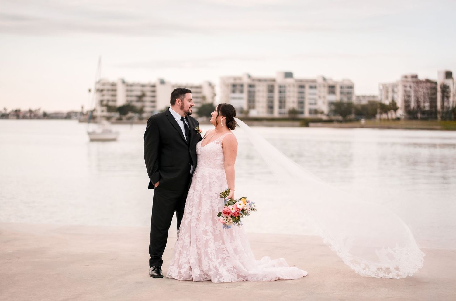 Newlyweds pose together on a beach with cityscape in the background at sunset.