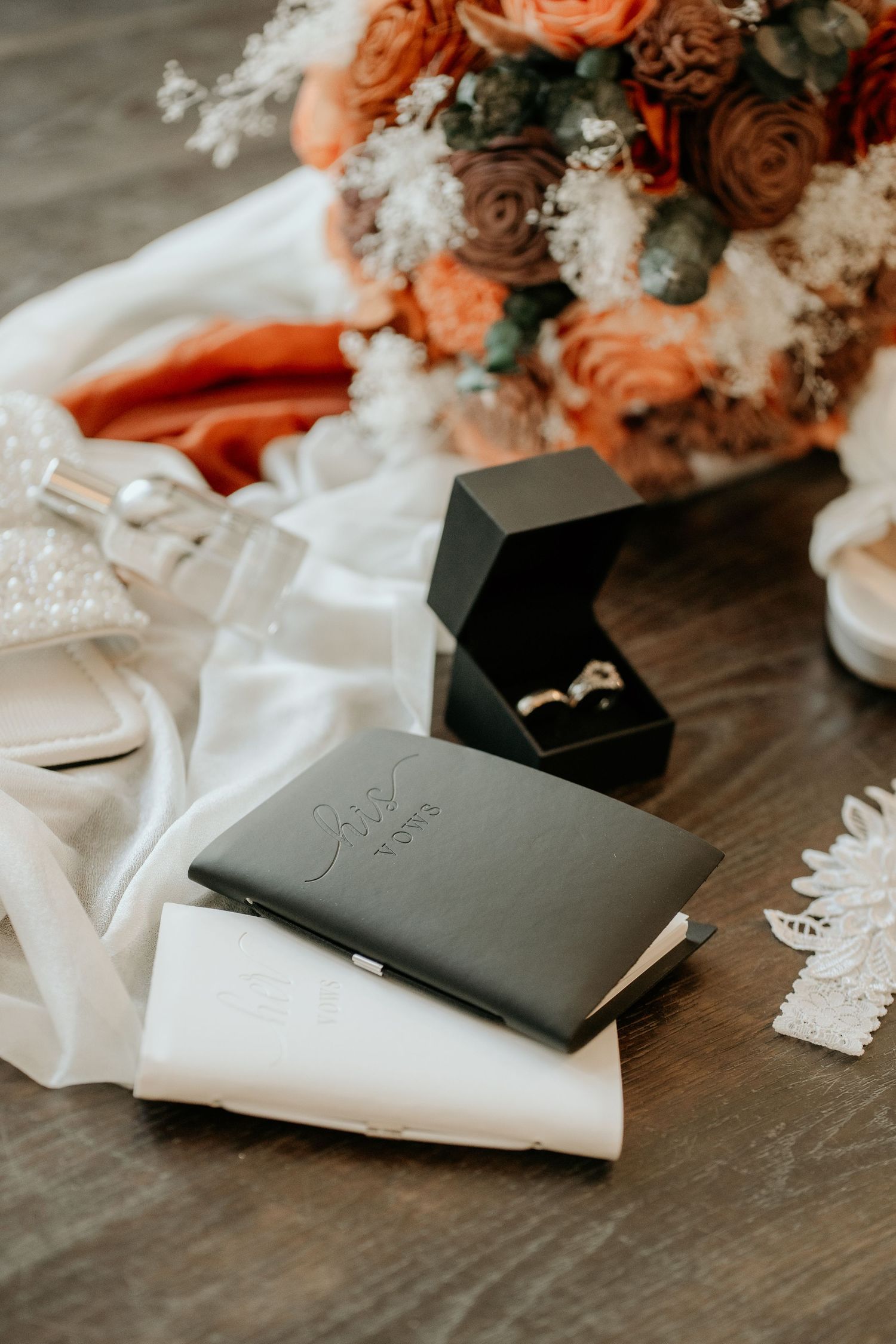 Wedding flat lay with two vow books, a black ring box holding gold bands, a rust floral bouquet, perfume bottle, and lace garter on wood.
