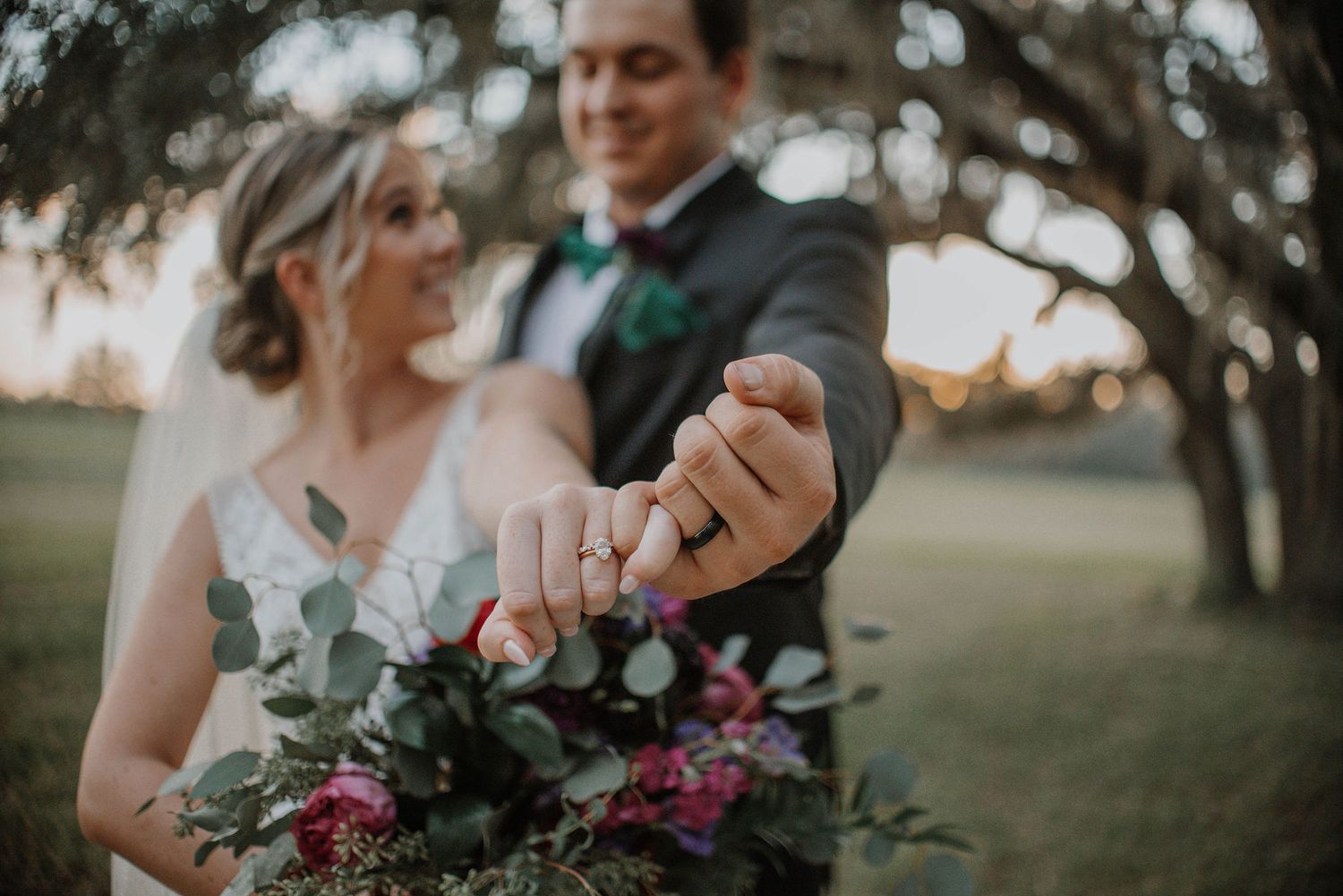 A couple shows off their wedding rings while holding a burgundy and green bouquet at sunset.