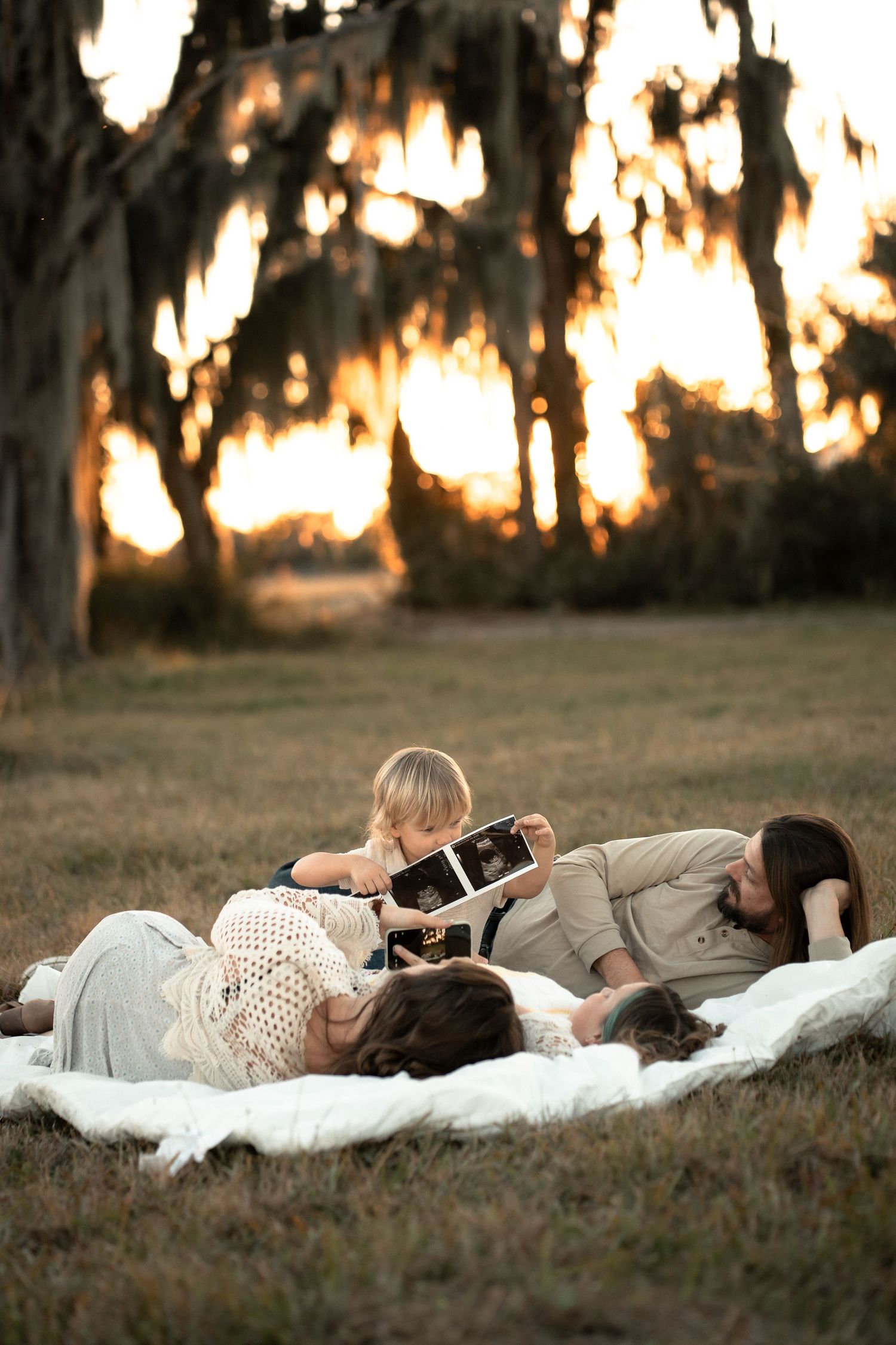 A peaceful family relaxes together on a blanket at sunset with Spanish moss hanging from trees in the background.