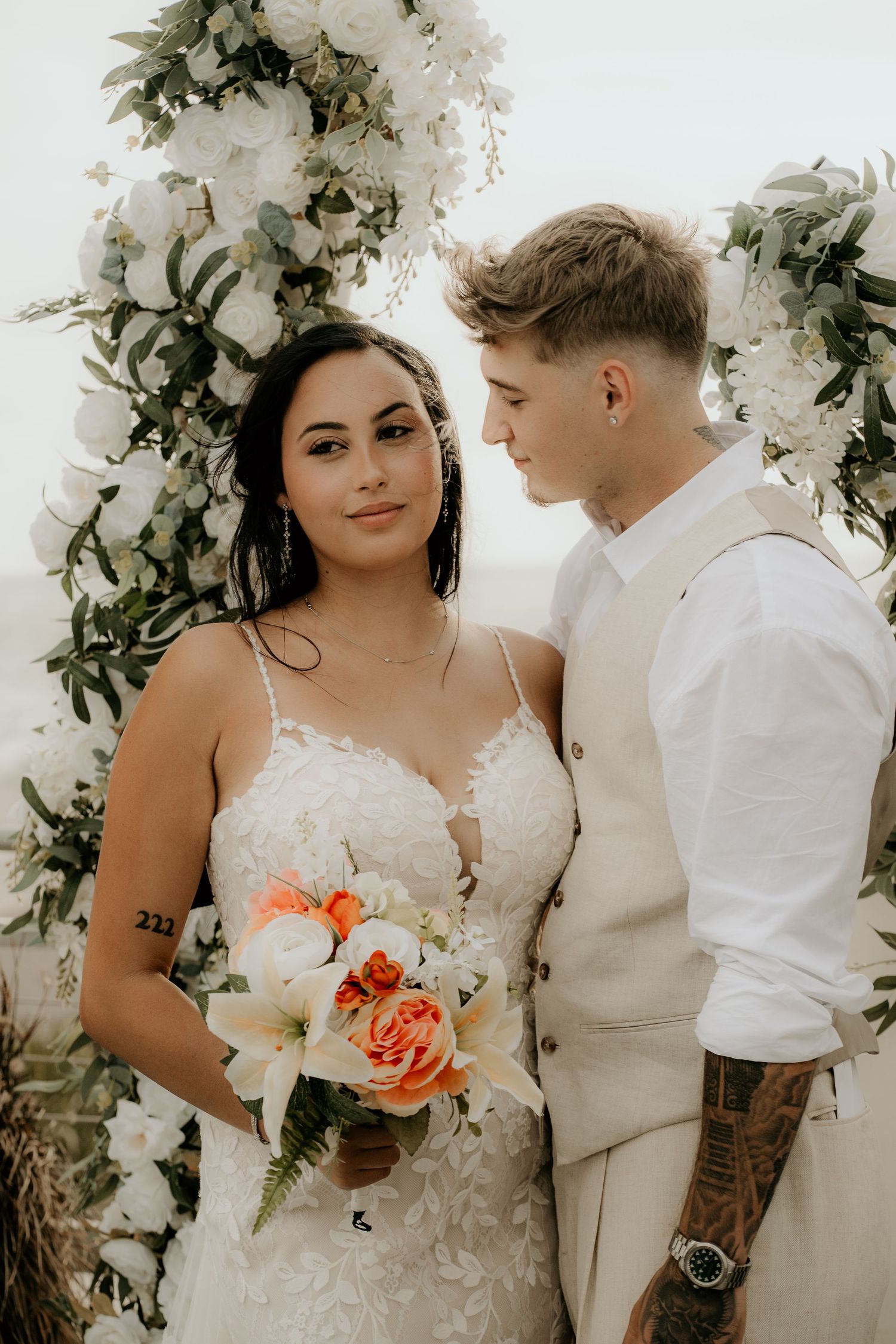 A couple poses against a white floral arch backdrop at their wedding ceremony.