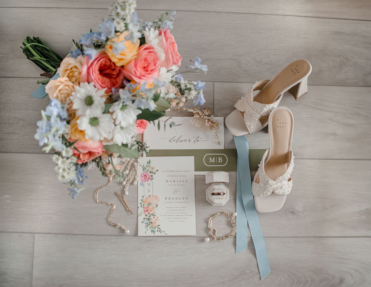 Wedding details flatlay with coral bouquet, invitation suite, and bridal shoes on wooden floor.