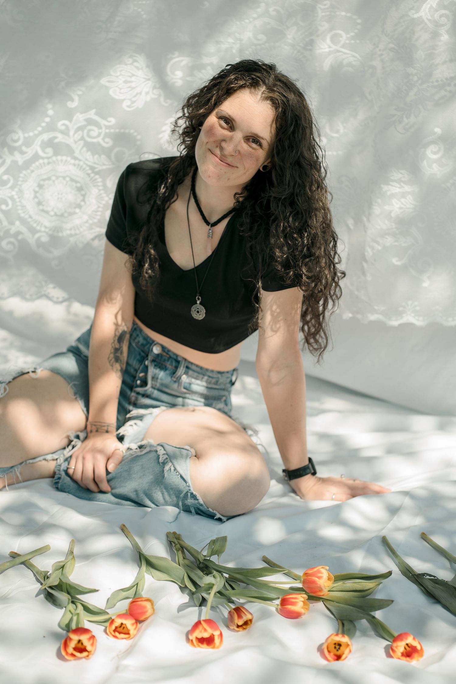 Woman with curly hair sits on white blanket with orange tulips in casual attire.