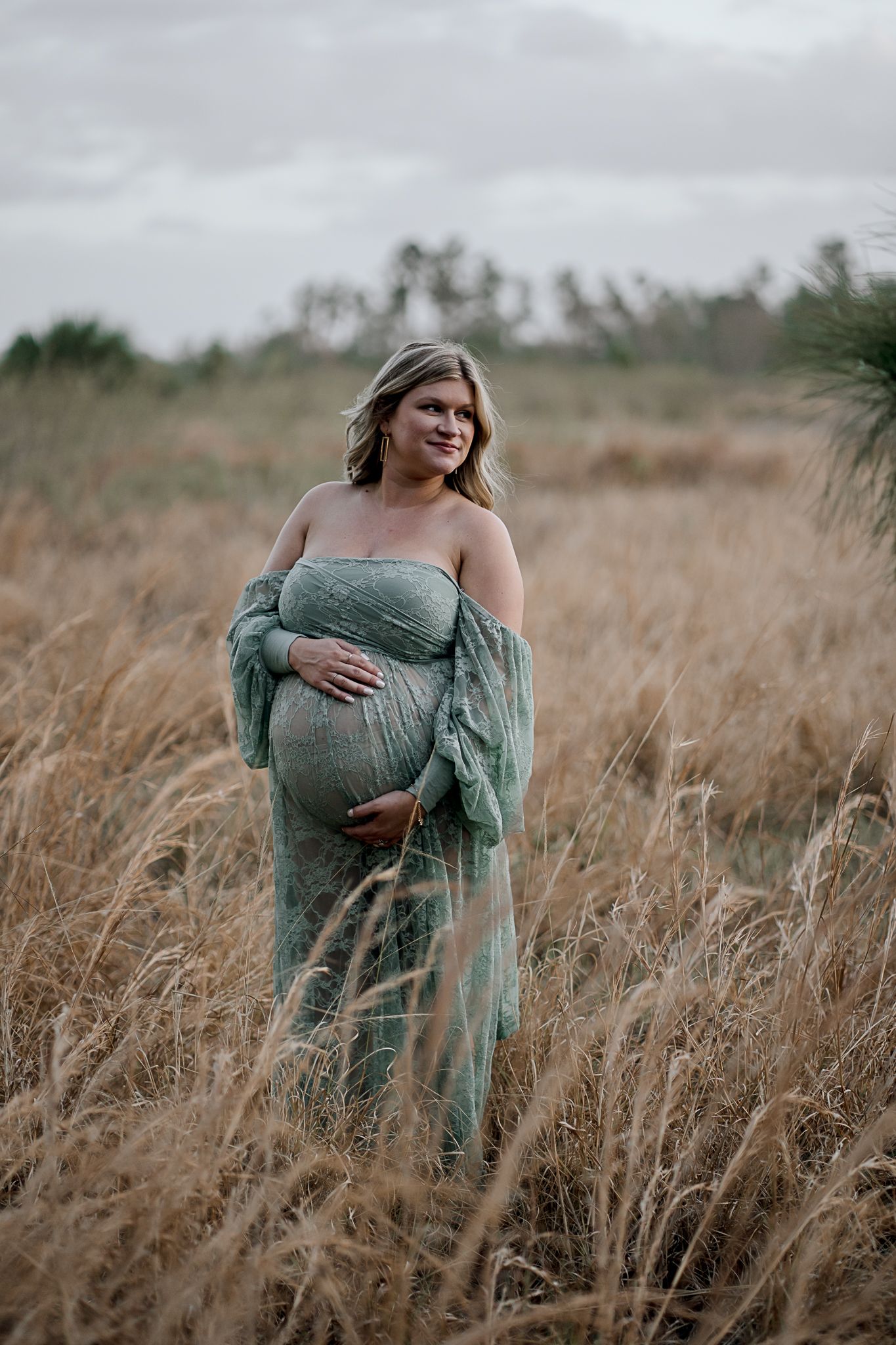 Maternity photo of woman in flowing sage green dress standing in tall golden grass at sunset.