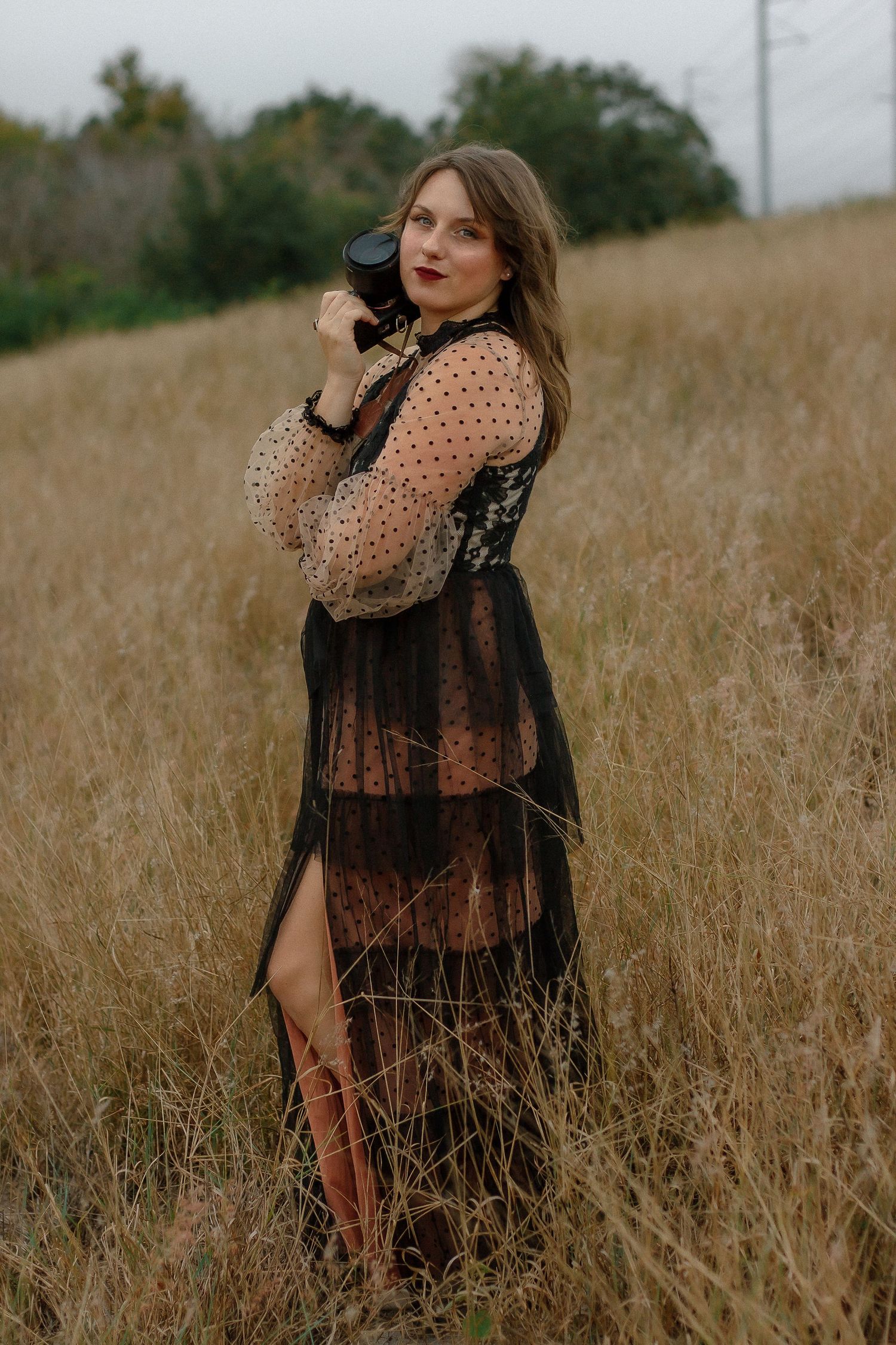 Woman in a black sheer polka dot dress with a high slit holds a camera while posing in a tall golden grass field at dusk.