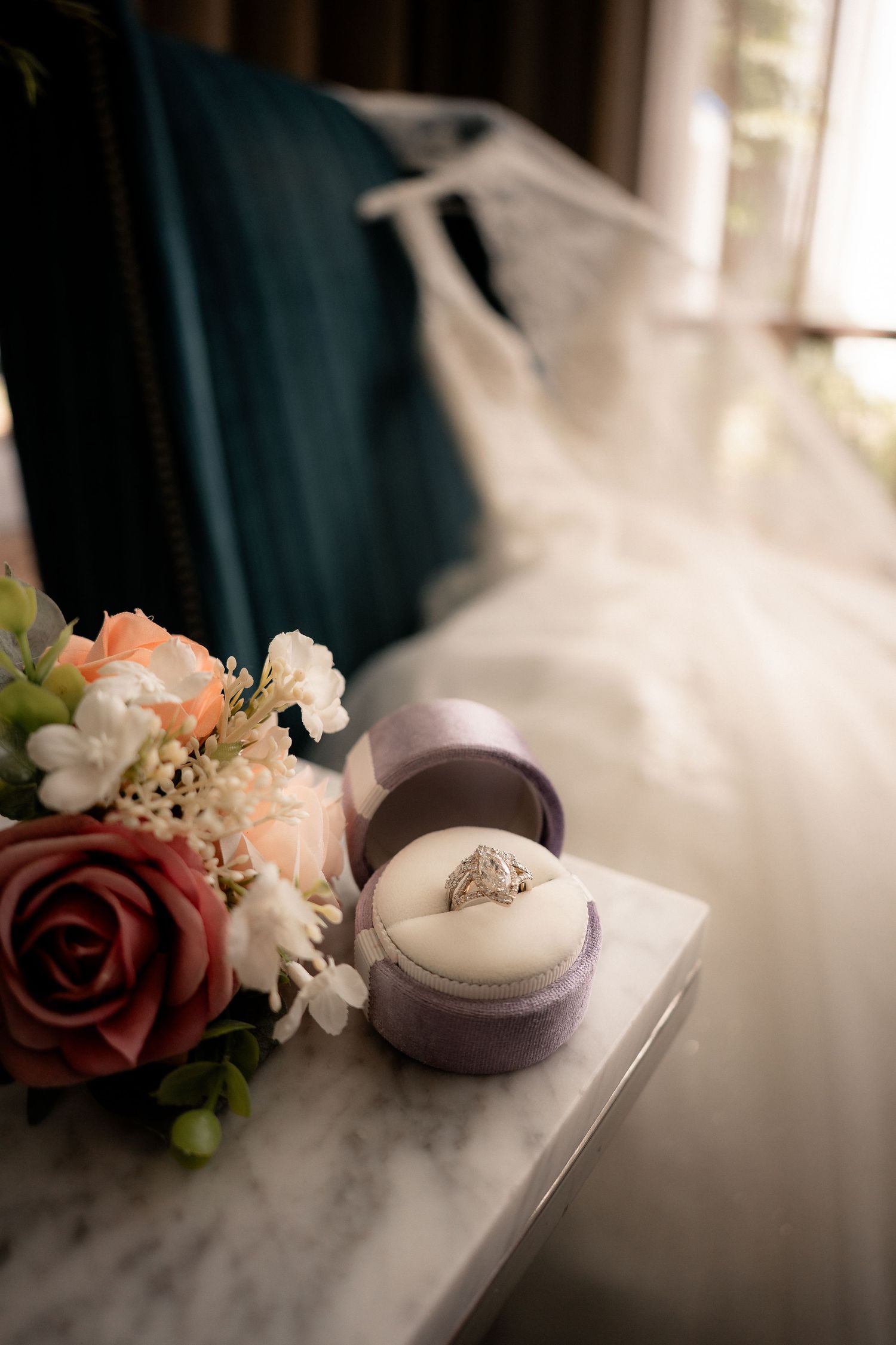 Diamond engagement ring in an open lavender velvet box on a marble table beside a rose bouquet, with a wedding dress in the background.