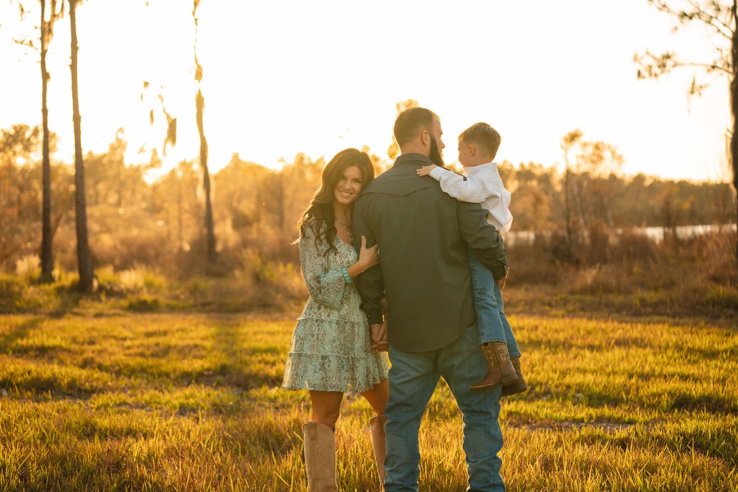 A family stands together in a golden sunlit field during sunset as warm light filters through the trees.