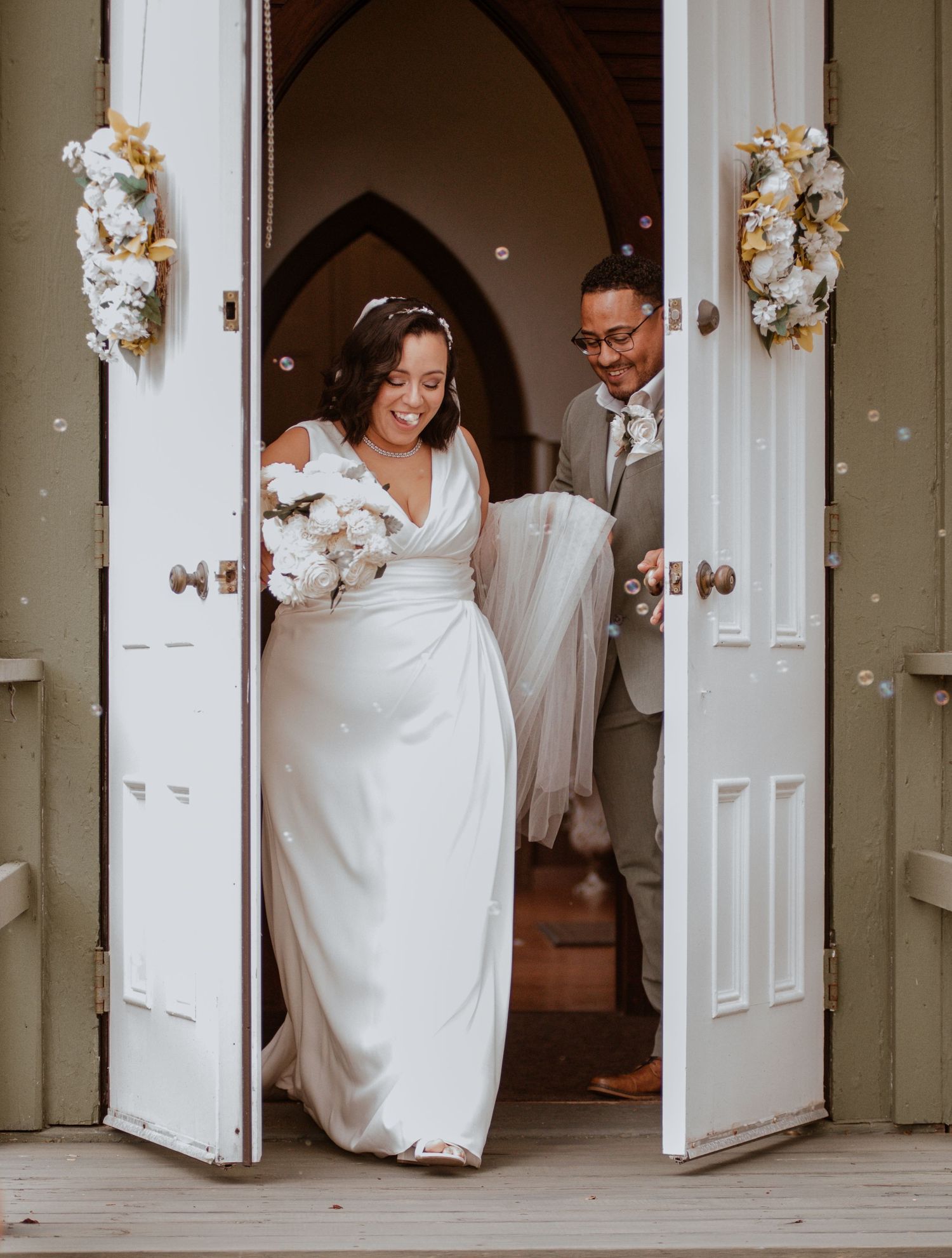 Wedding couple exits through ornate church doors decorated with white floral arrangements.