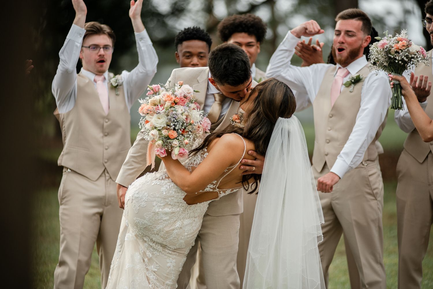 Wedding party celebrates with raised arms as newlyweds share a kiss outdoors.
